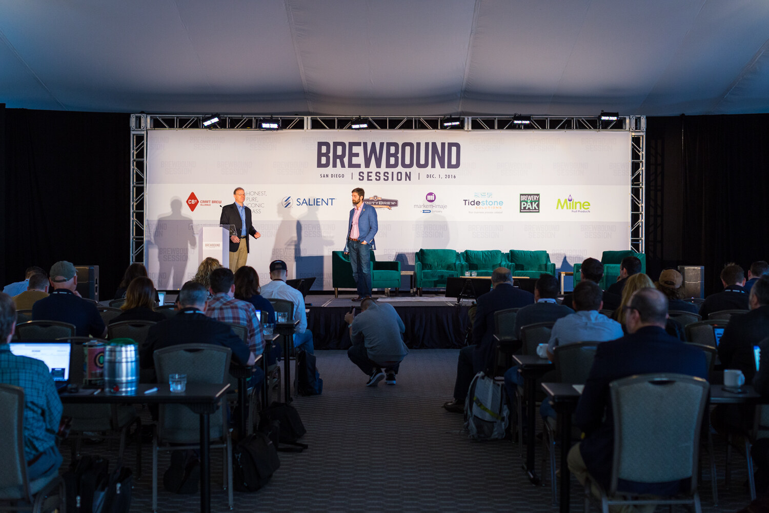 Wide view of the darkened ballroom with rows of attendees facing a lit stage, a banner reading Brewbound Session San Diego, Dec. 1, 2016 and two people standing on stage.
