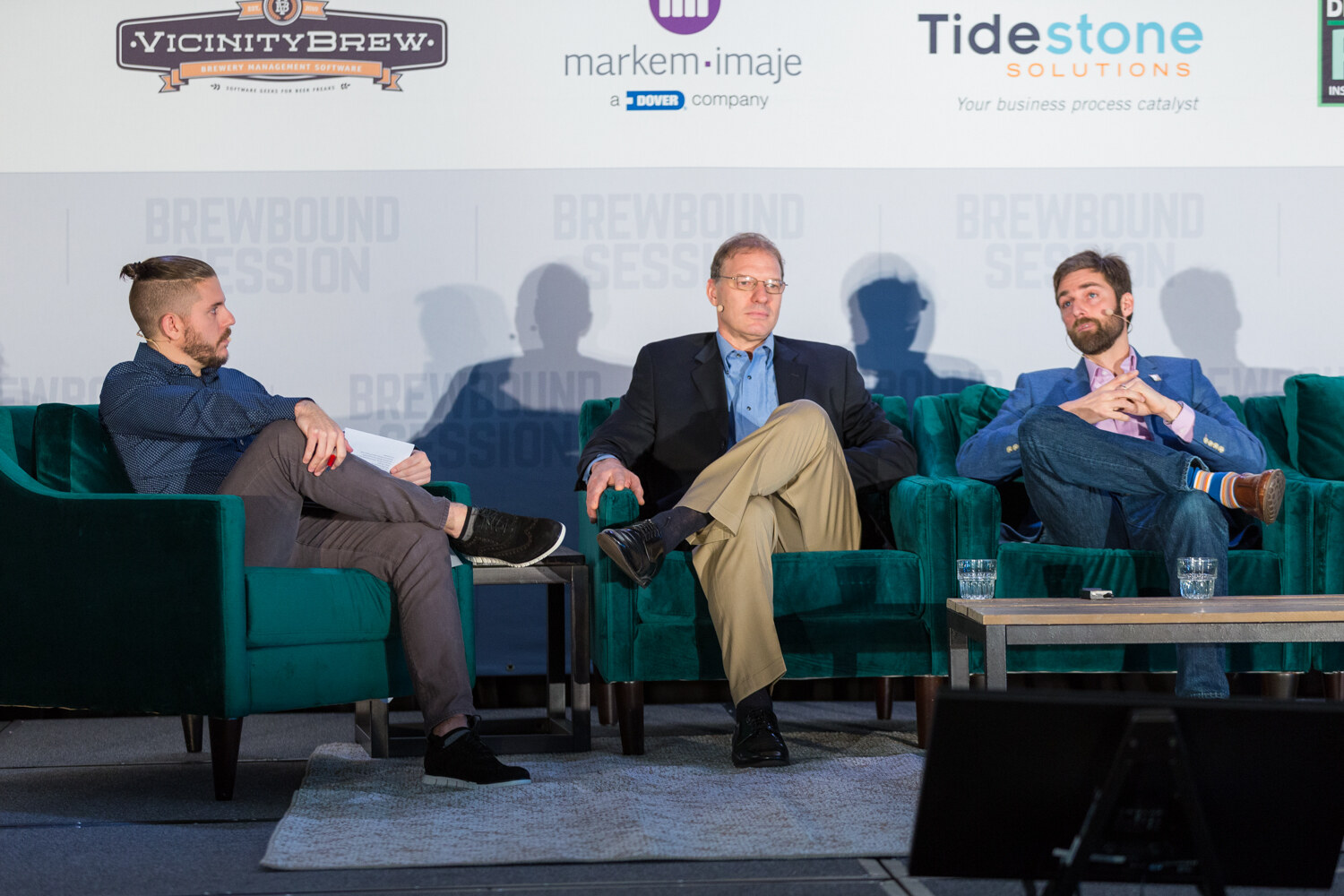 Wide view of three men seated on green sofas on stage under logos for Vicinity Brew, Markem-Imaje and Tidestone Solutions.