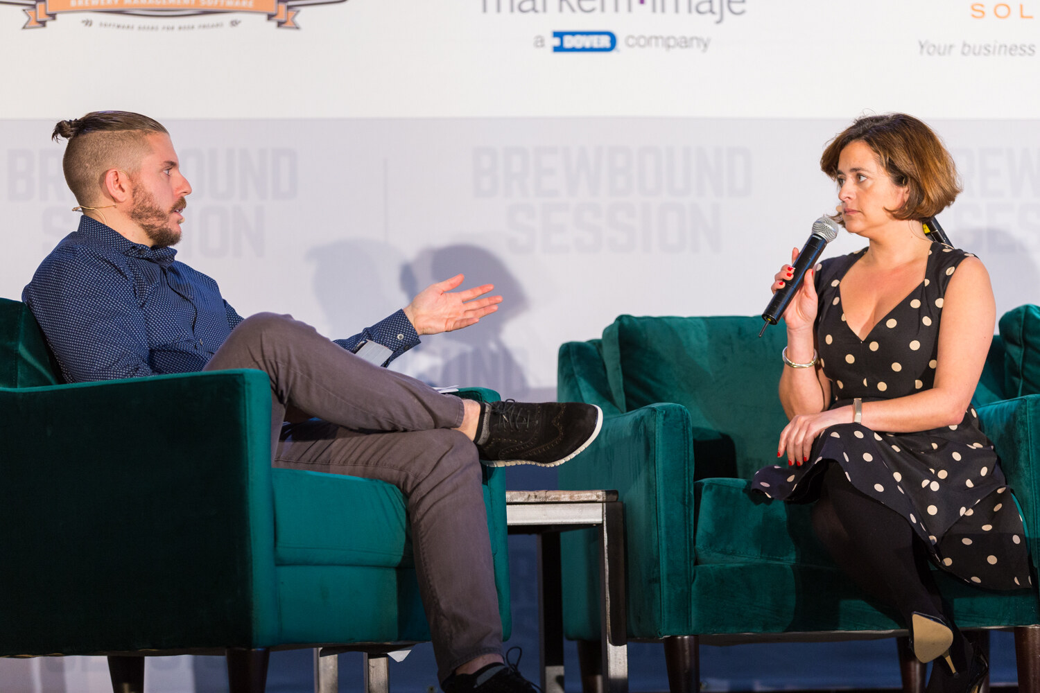 Bearded man in a navy shirt gesturing at left and a woman in a polka-dot dress holding a microphone at right, both in green velvet armchairs.