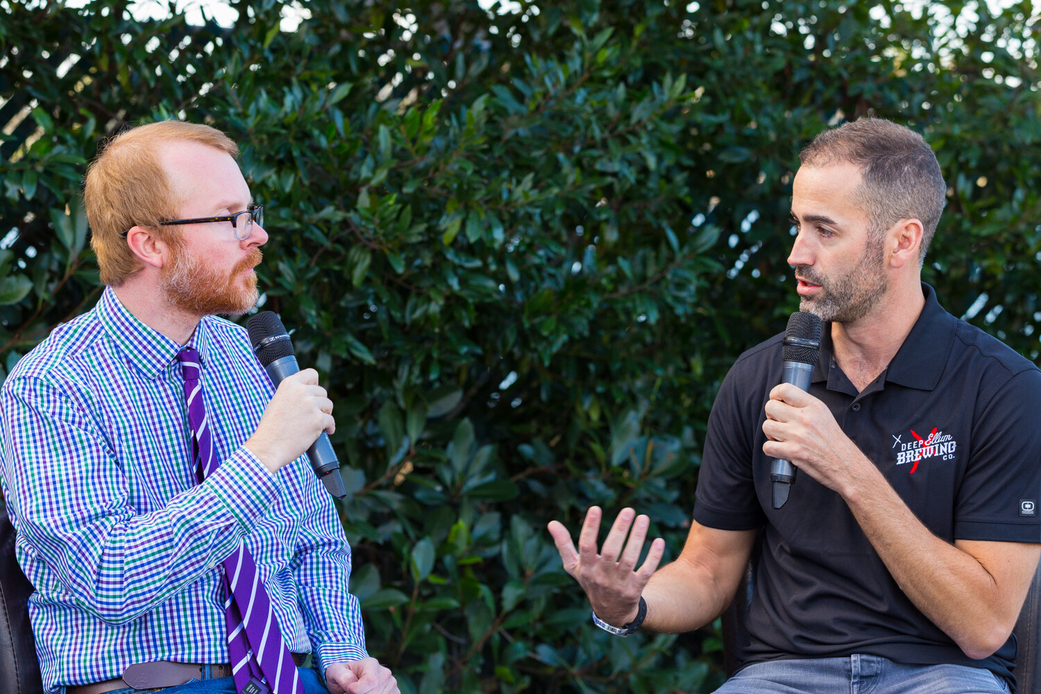 Man in a checked shirt and purple tie at left and a man in a black Deep Ellum Brewing polo gesturing at right, both holding microphones in front of green foliage.