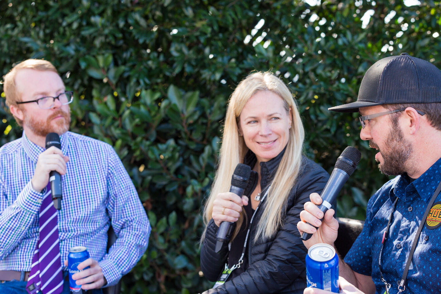 Woman with long blond hair smiling between a man in a checked shirt and a man in a black cap, all holding microphones in front of a hedge, with blue beer cans in the foreground.
