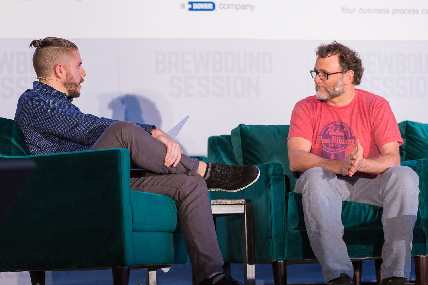 Man in a navy shirt with notes on his lap facing a man in glasses and a red Pabst Blue Ribbon T-shirt, seated on green velvet armchairs.