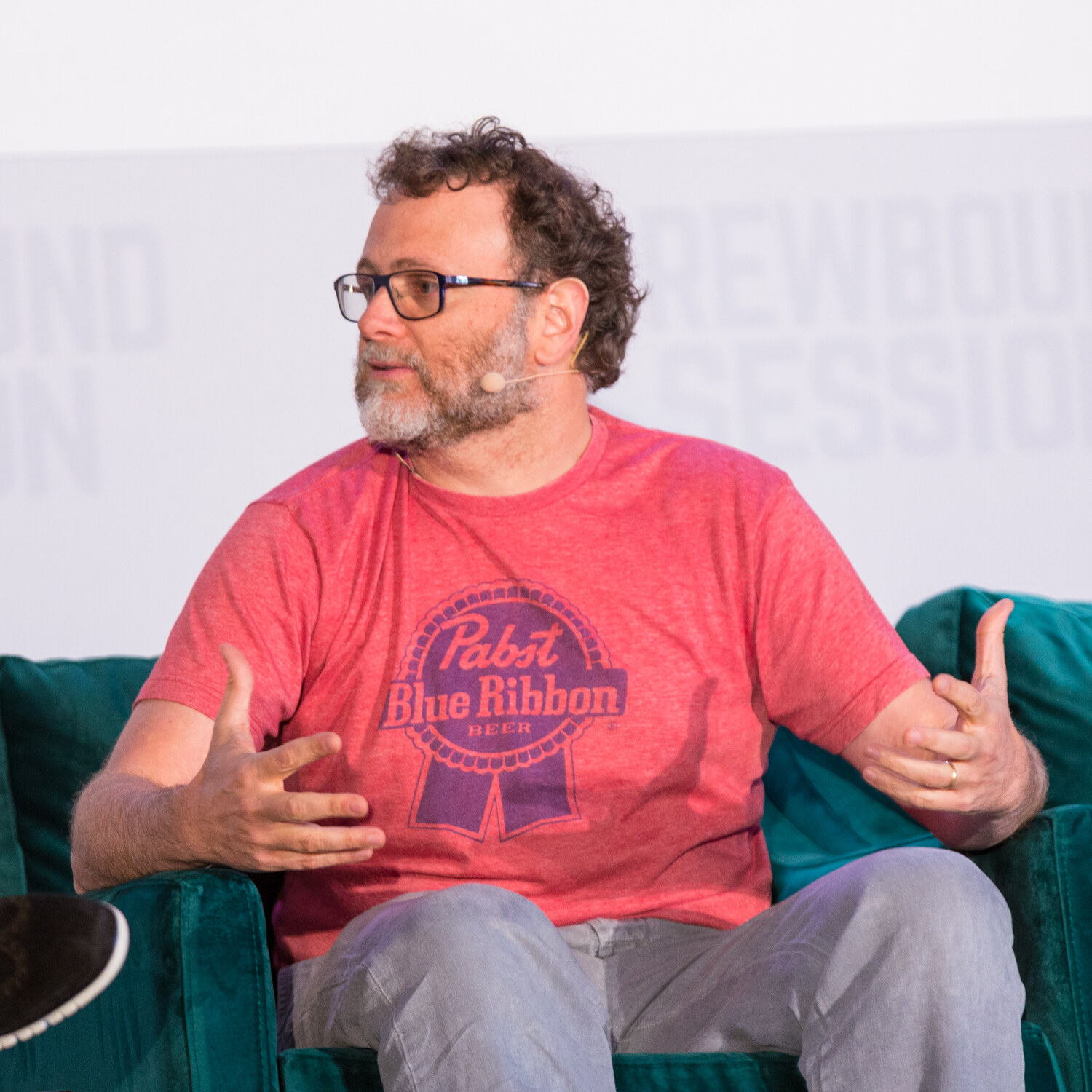 Close view of a bearded man in glasses with a headset microphone and a red T-shirt with the Pabst Blue Ribbon logo, seated on a green sofa.