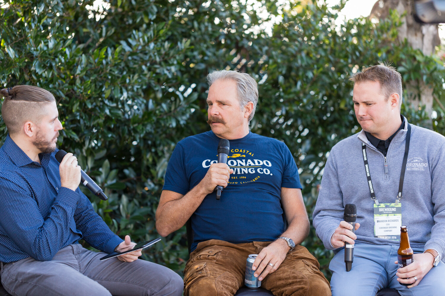 Man in a navy Coronado Brewing Co. T-shirt speaking into a microphone between an interviewer in a blue shirt and a man in a gray quarter-zip with a Brewbound Session badge, in front of a hedge.