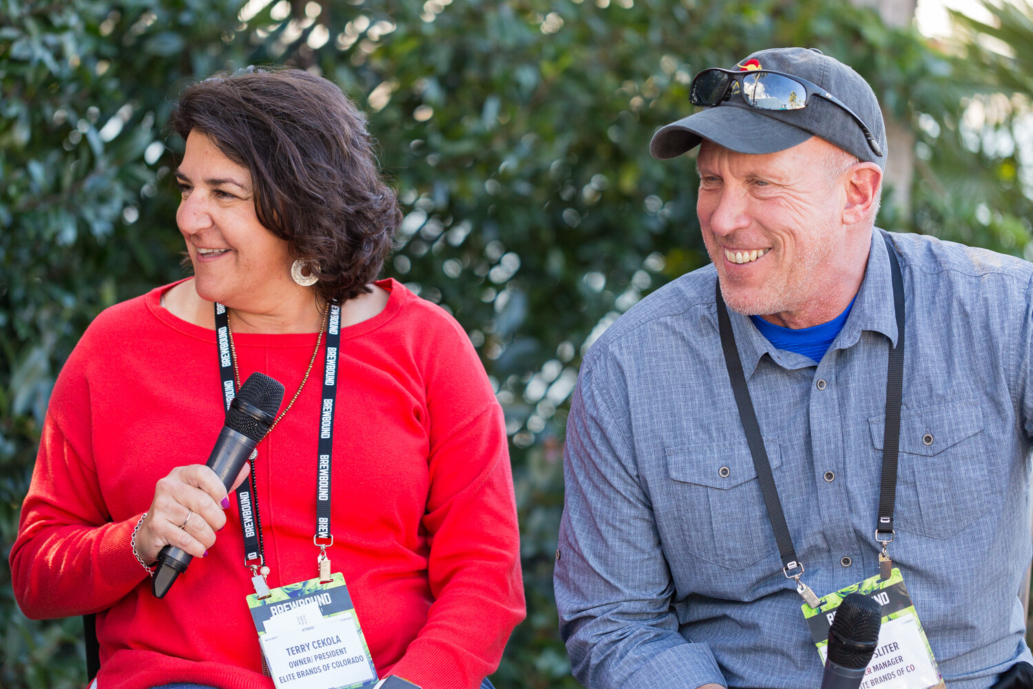 Woman in a red sweater holding a microphone and a man in a gray cap and denim shirt smiling beside her, both with Brewbound lanyards in front of foliage.