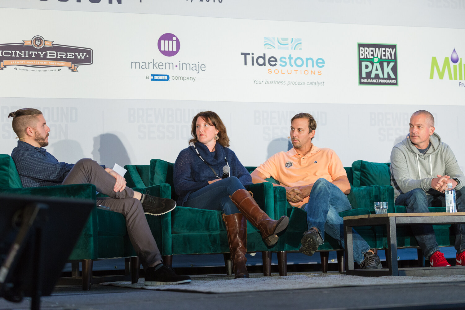 Wide view of four people seated on green sofas on stage, a bearded man in a navy shirt at left, a woman in a navy sweater, a man in a peach polo and a man in a gray hoodie, under a row of sponsor logos.