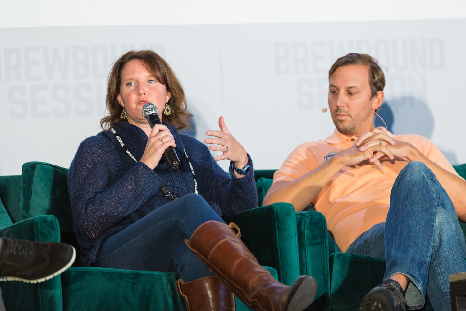 Woman in a navy sweater with a lanyard and gold earrings holding a microphone and gesturing, with a man in a peach polo seated beside her on a green sofa.