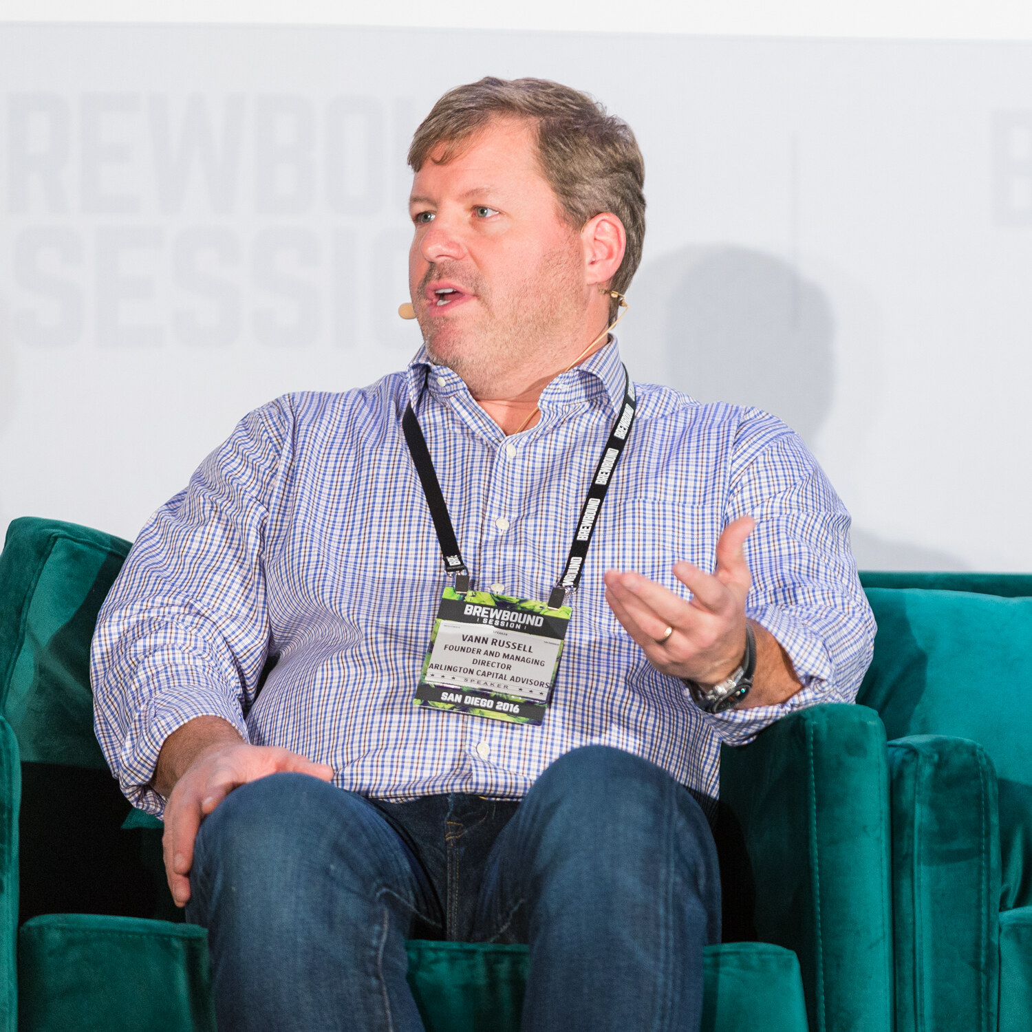 Man in a blue checked shirt and jeans wearing a Brewbound Session San Diego 2016 speaker badge, gesturing from a green velvet armchair.