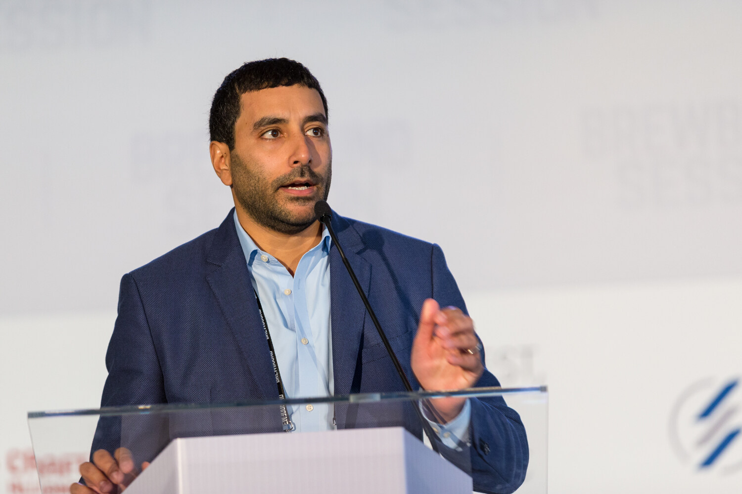 Man in a navy blazer and light blue shirt with a lanyard speaking into a microphone at a clear lectern, with partial sponsor logos behind him.