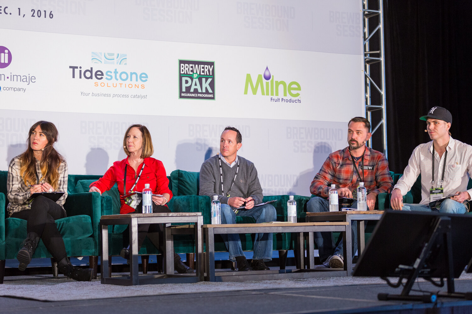 Wide view of five people seated side by side on green sofas holding clipboards and bottled water, under a backdrop dated Dec. 1, 2016.