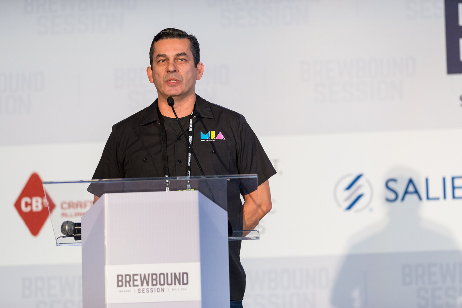 Man in a black work shirt with an MIA Beer Company logo standing behind a lectern labeled Brewbound Session, with a Craft Brew Alliance logo and Salient logo behind.