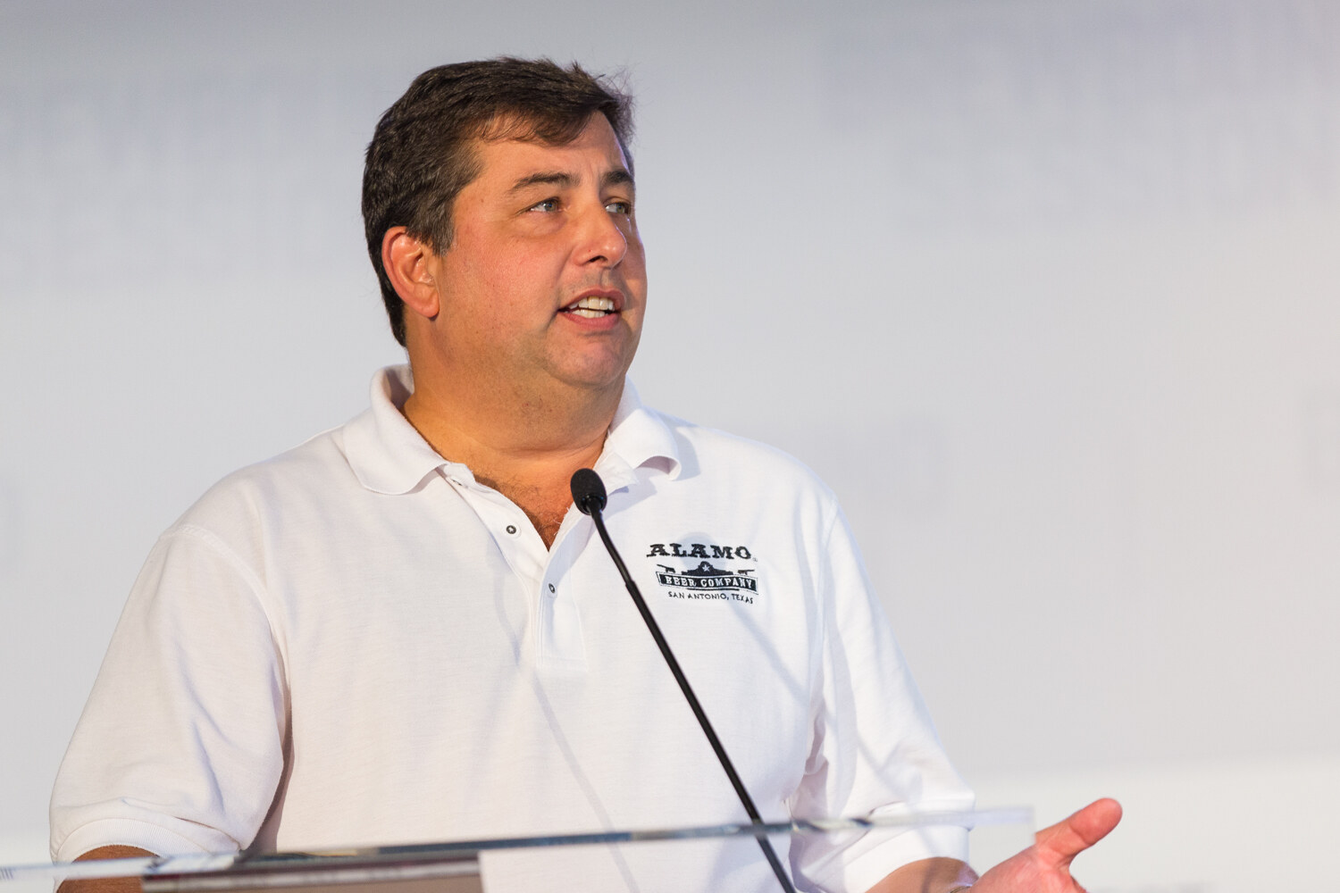 Man in a white polo shirt with an Alamo Beer Company, San Antonio, Texas logo speaking at a clear lectern.
