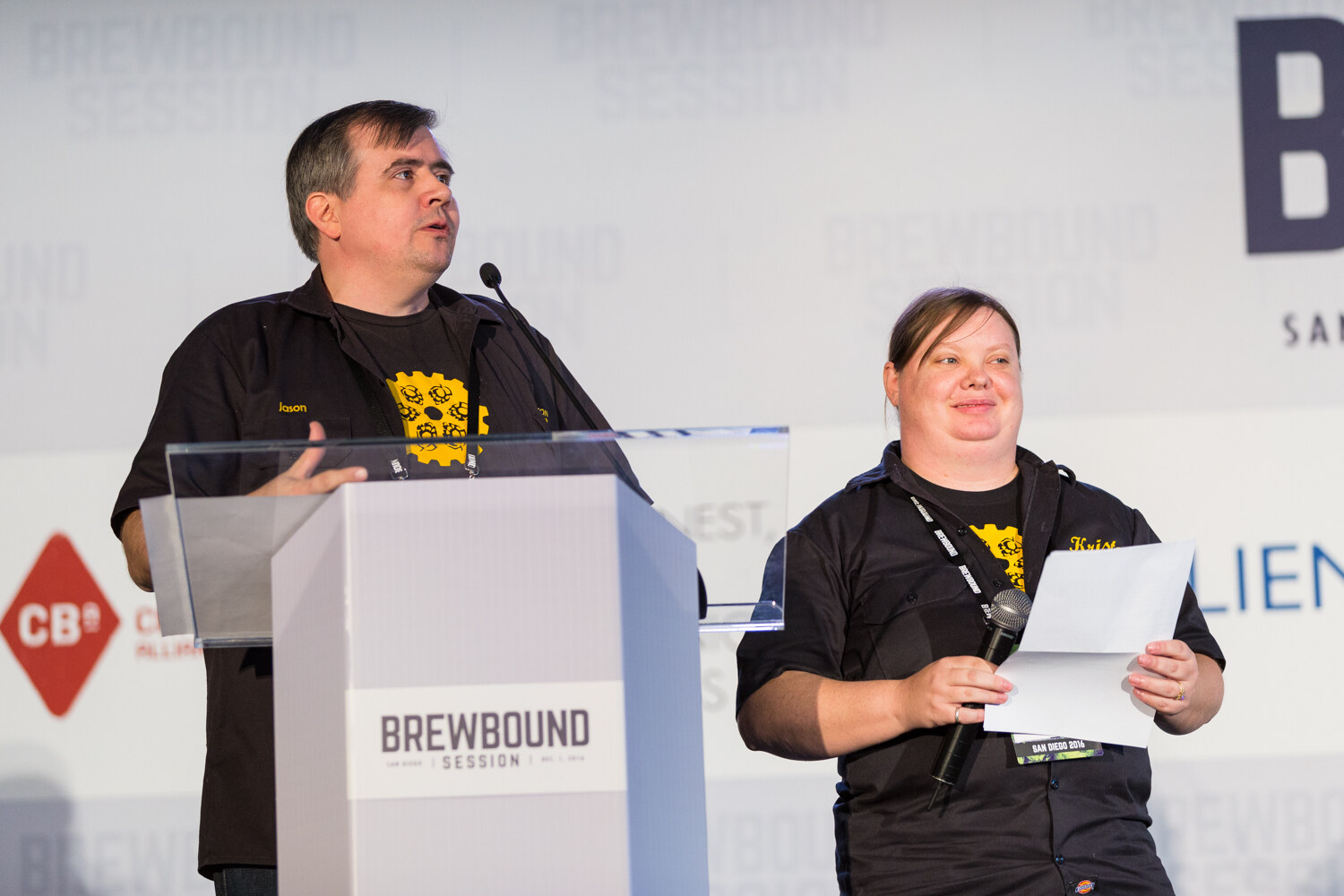 Man in a black shirt with a yellow gear logo speaking at a lectern and a woman beside him holding a microphone and a sheet of paper, with a Craft Brew Alliance logo in the background.