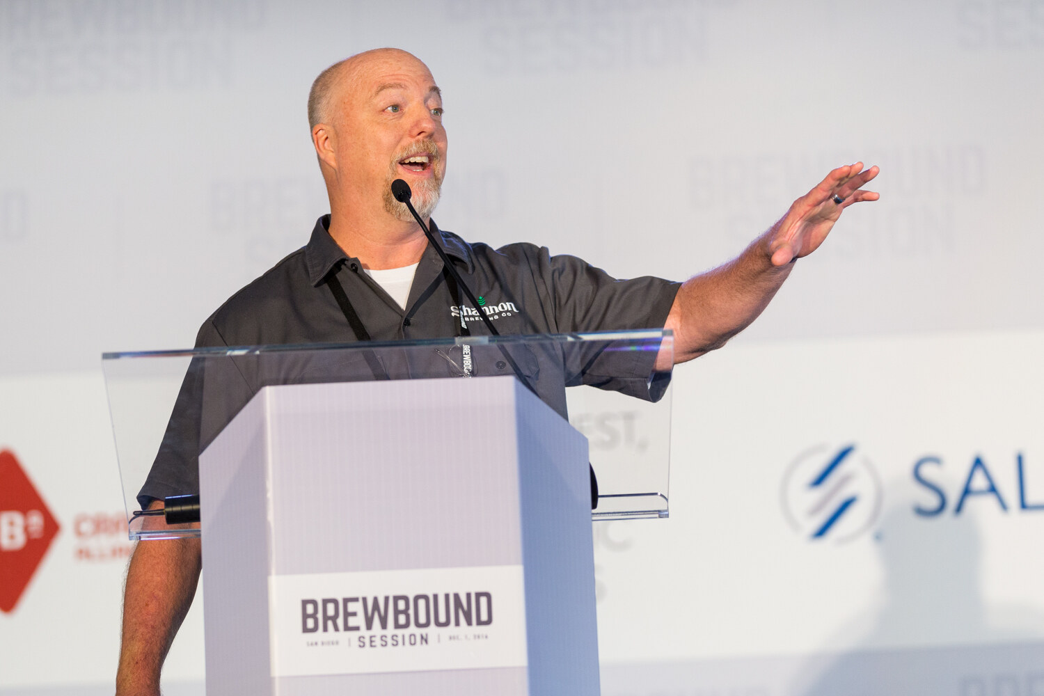 Man in a gray Shannon Brewing Co. shirt with one arm outstretched behind a Brewbound Session lectern, with Craft Brew Alliance and Salient logos behind him.