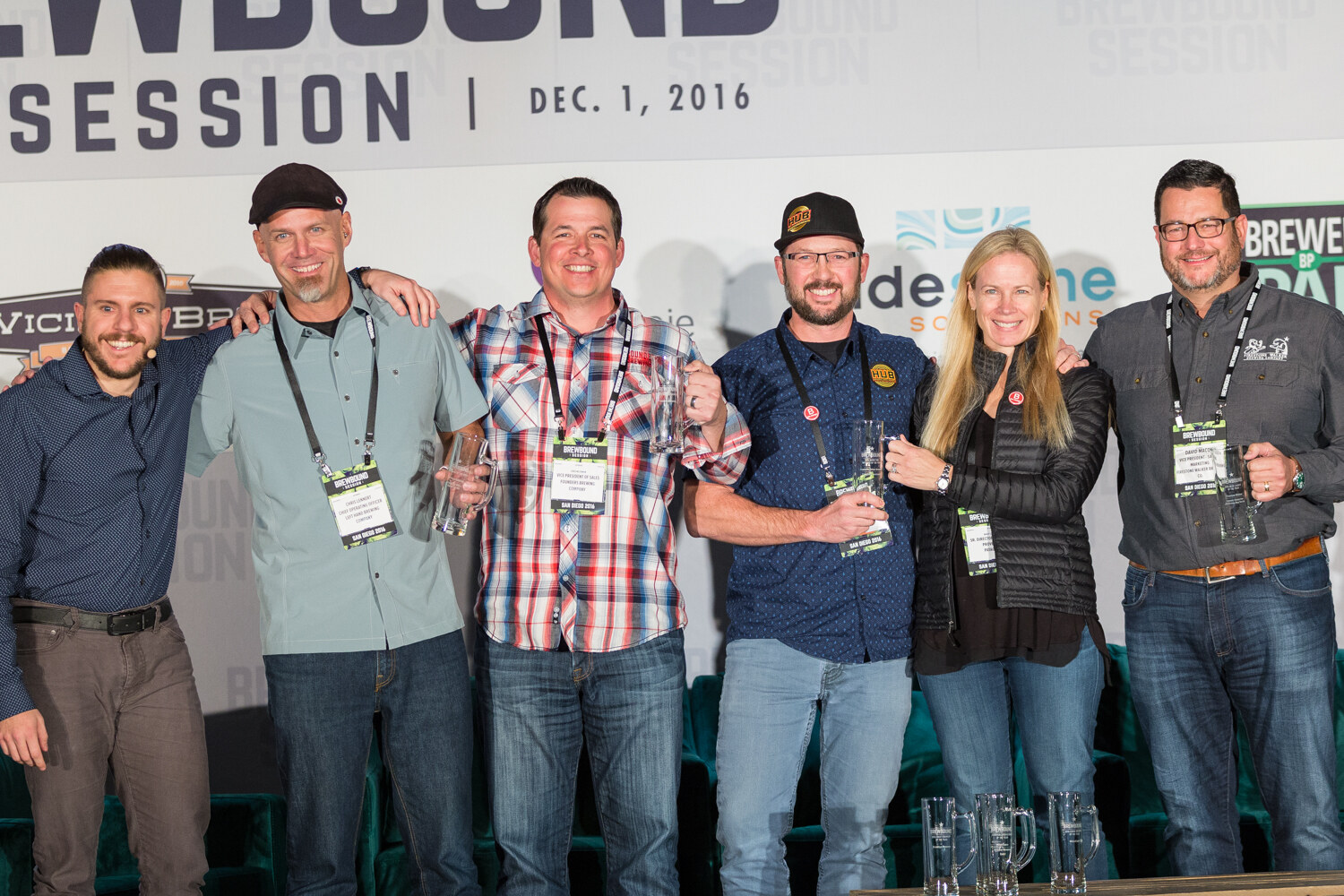 Six people with Brewbound Session badges standing in a row on stage, several holding glass beer mugs, with a Dec. 1, 2016 banner above.