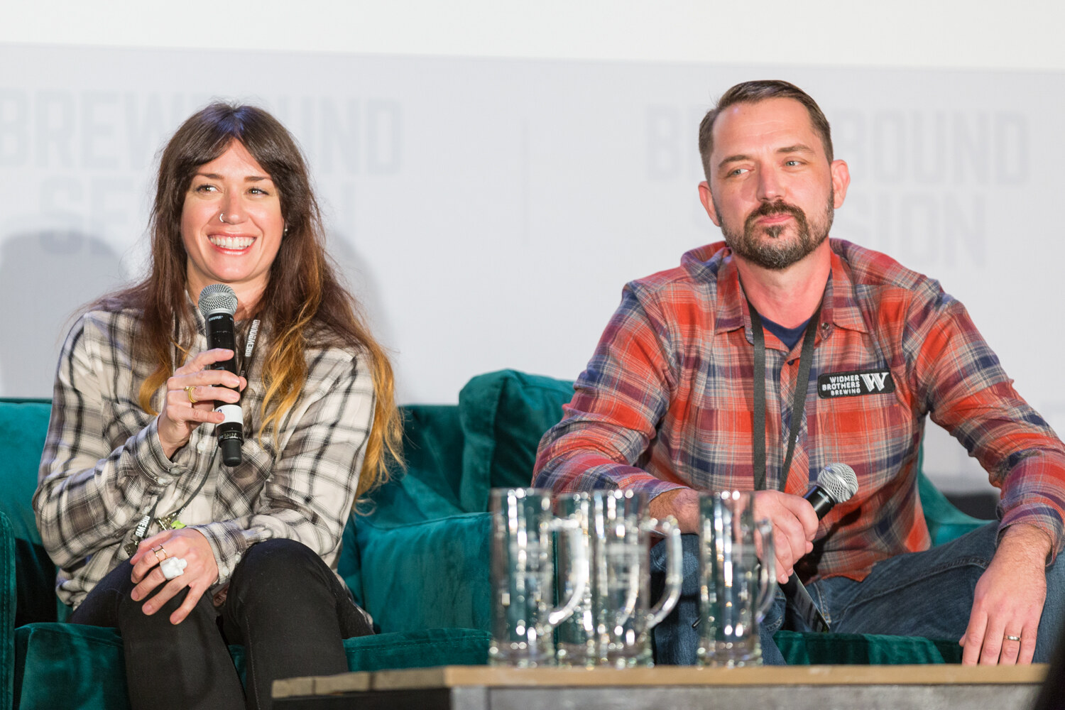 Woman in a plaid shirt smiling and holding a microphone beside a bearded man in a red plaid Widmer Brothers Brewing shirt with beer mugs on the table in front of them.