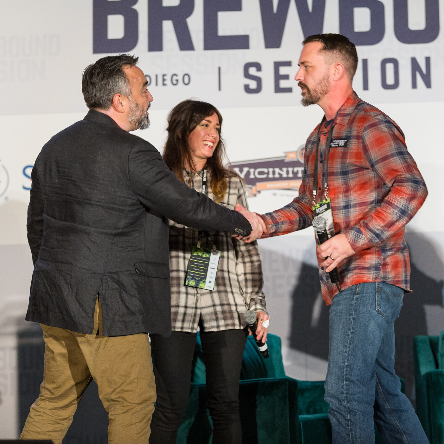 Man in a charcoal blazer with his back to the camera shaking hands with a man in a red plaid Widmer Brothers Brewing shirt, with a smiling woman between them under the Brewbound Session banner.