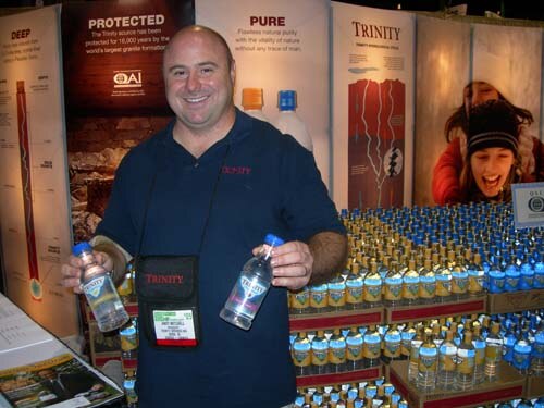 A smiling bald man in a dark Trinity shirt holds two water bottles in front of a display of Trinity bottles and banners reading Protected, Pure and Deep.