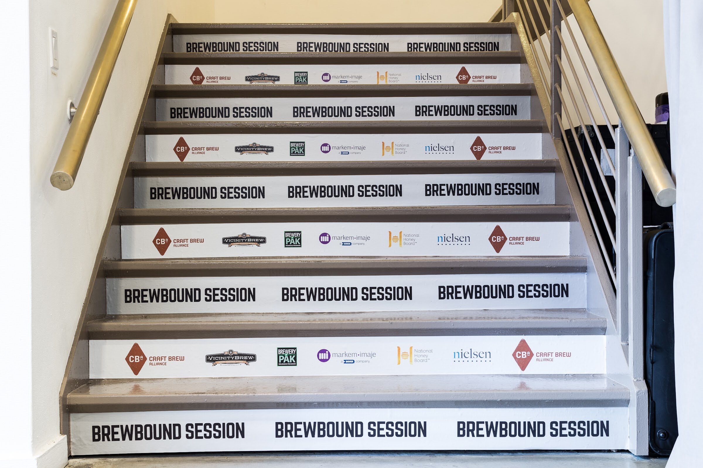 A staircase with brass handrails whose risers read Brewbound Session alternating with rows of sponsor logos including Craft Brew Alliance, Brewery Pak and Nielsen.