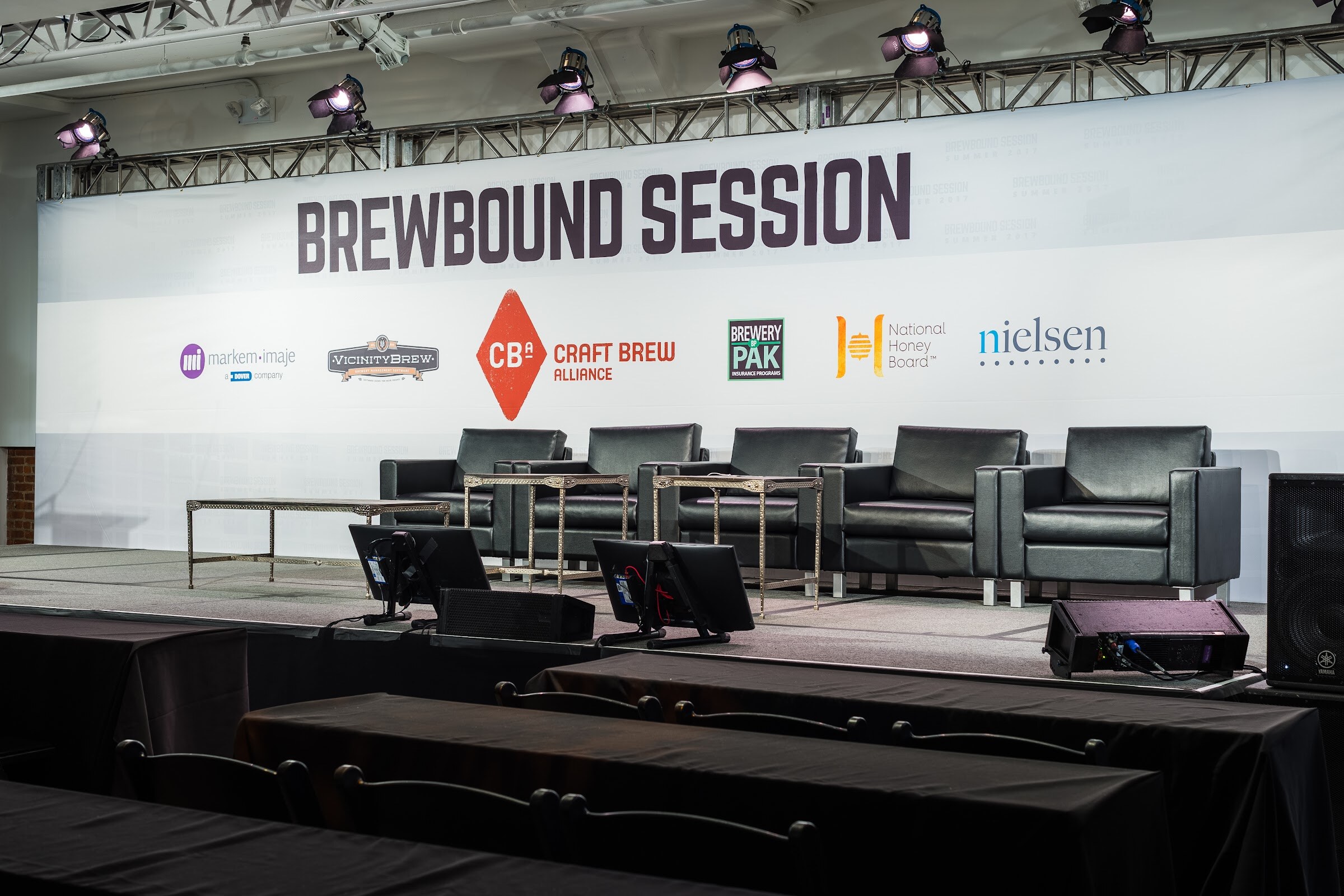 A stage with five black leather armchairs and side tables in front of a Brewbound Session banner with Markem-Imaje, Vicinity Brew, Craft Brew Alliance, Brewery Pak, National Honey Board and Nielsen logos.