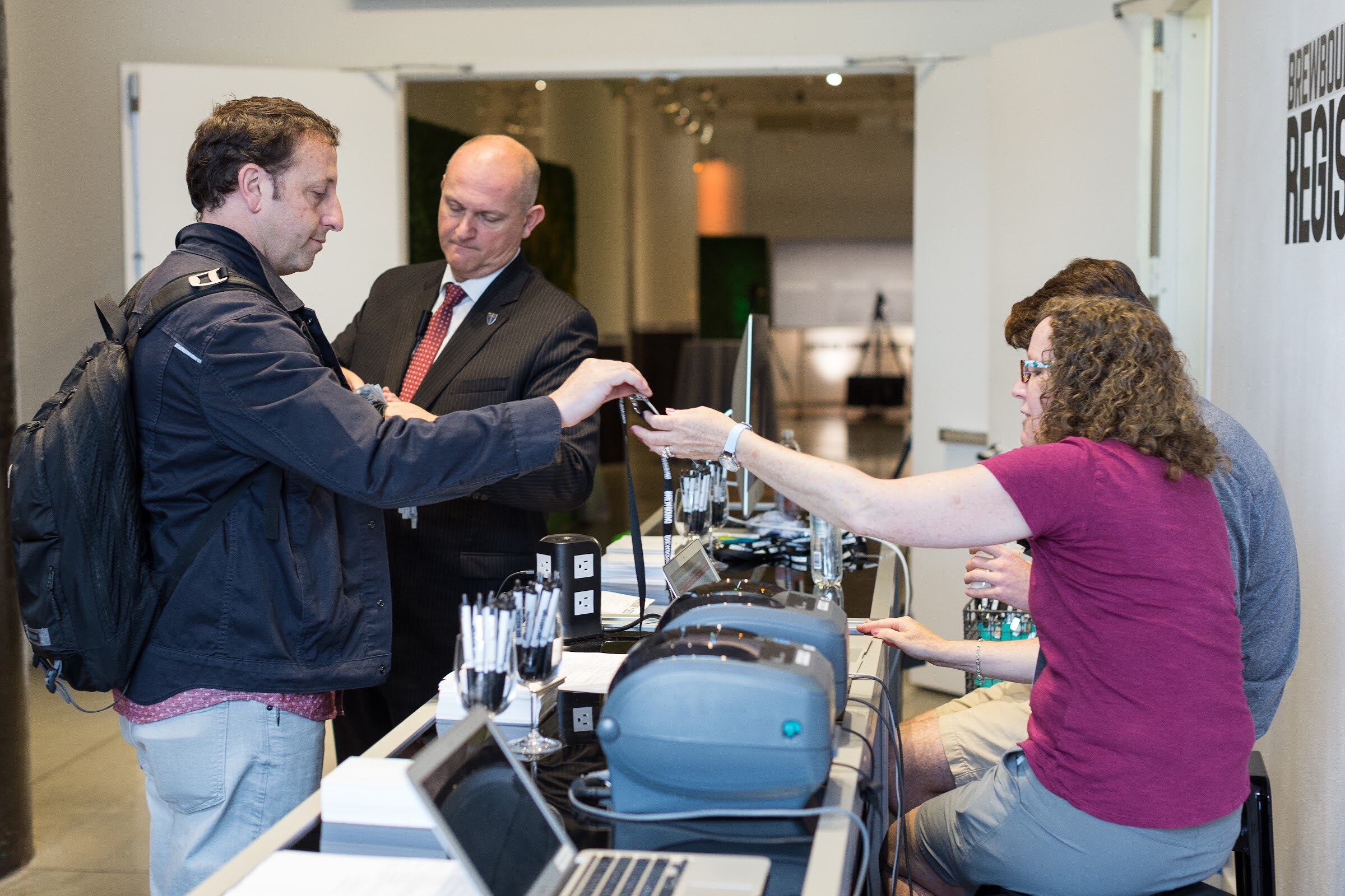 A man with a black backpack takes a badge on a lanyard from a woman at a registration desk with label printers and a laptop while a man in a suit stands behind him.