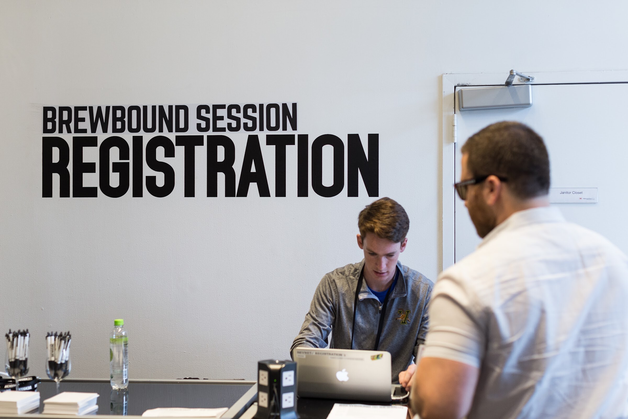 A young man in a gray quarter-zip works on a MacBook at a desk below a wall sign reading Brewbound Session Registration while an attendee in a white shirt stands at the counter.