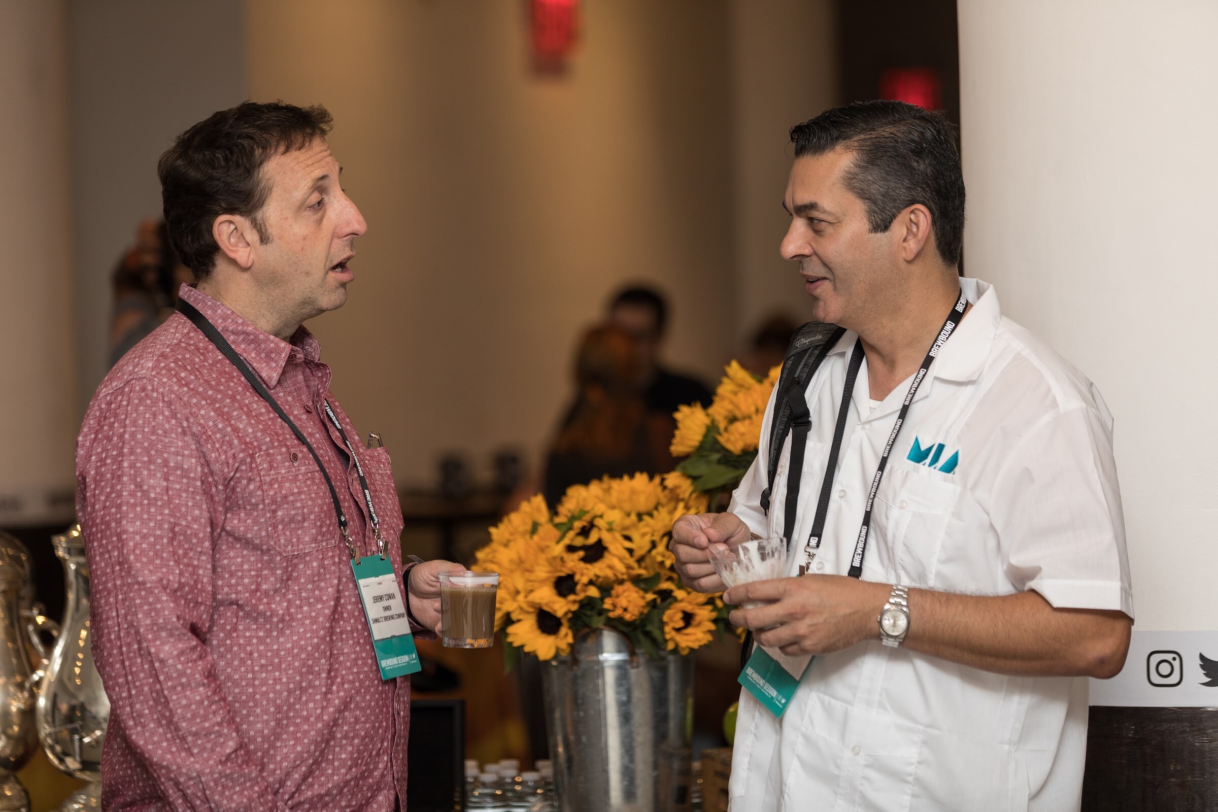 A man in a pink patterned shirt and a man in a white shirt with an M.I.A. logo talk while holding cups next to a vase of sunflowers.