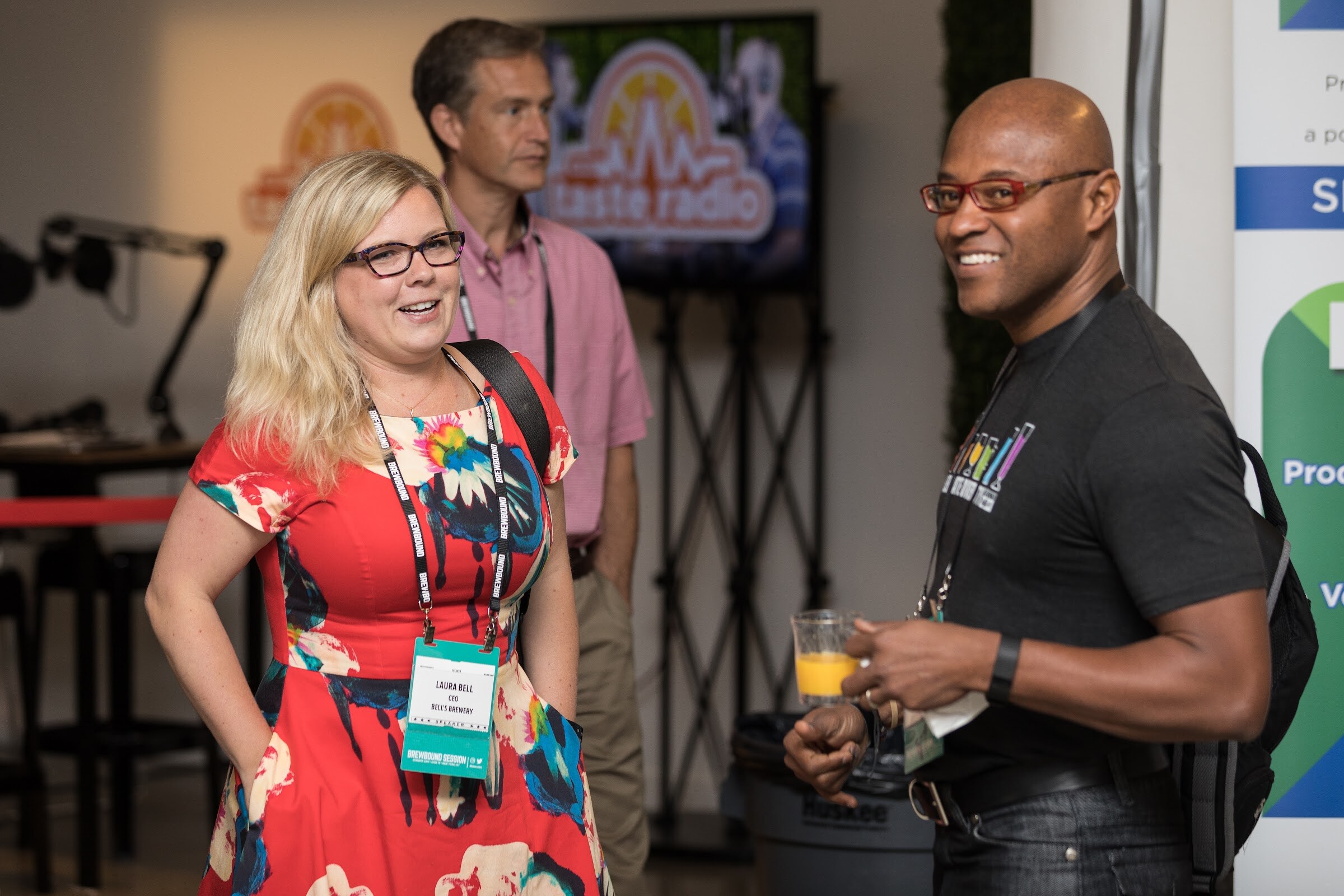 A woman in a red floral dress and a smiling man with red-framed glasses hold drinks and talk in front of a Taste Radio screen and microphone arm.