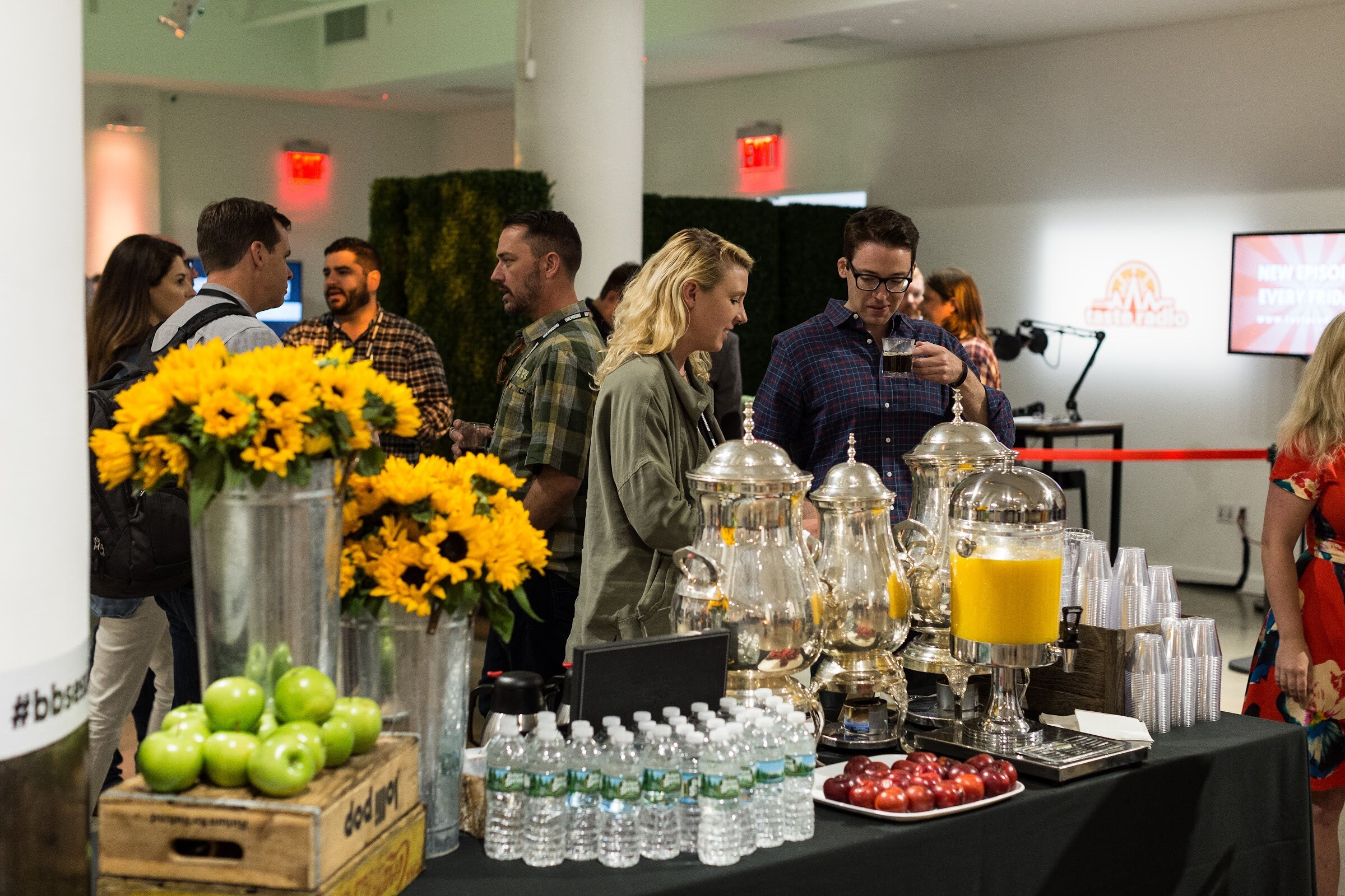 Attendees stand around a black-draped table holding silver coffee urns, an orange juice dispenser, bottled water, a crate of green apples and vases of sunflowers, with a Taste Radio screen in the background.