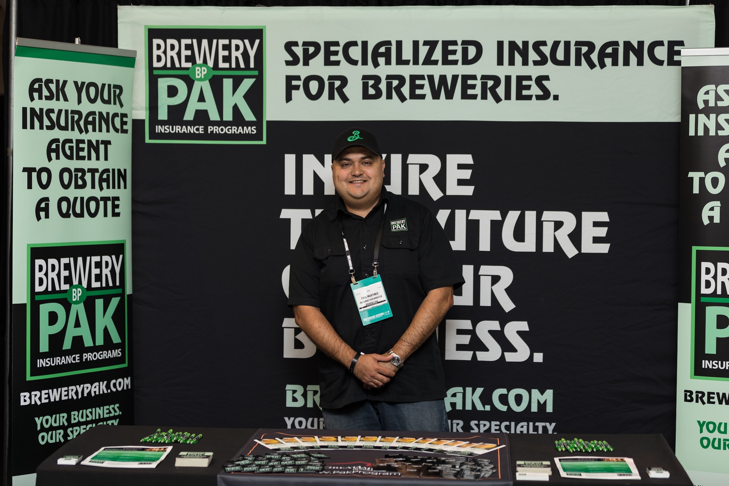 A man in a black Brewery Pak shirt and cap stands behind a table in front of a black and green Brewery Pak backdrop reading Specialized Insurance for Breweries.