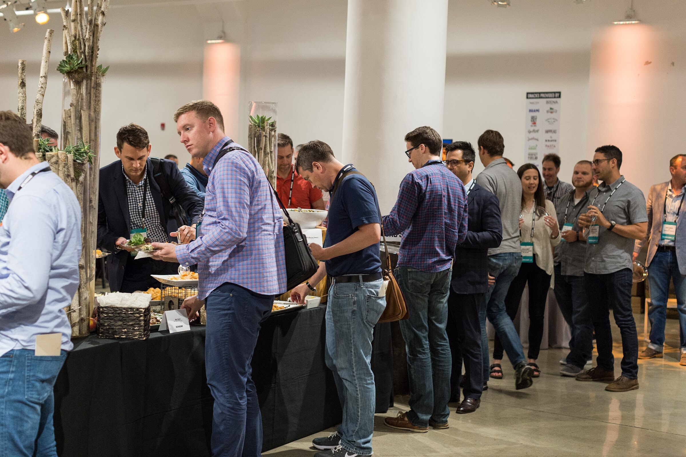 A line of attendees with badge lanyards fill plates at a black-draped buffet table in a white room with birch-branch decor.