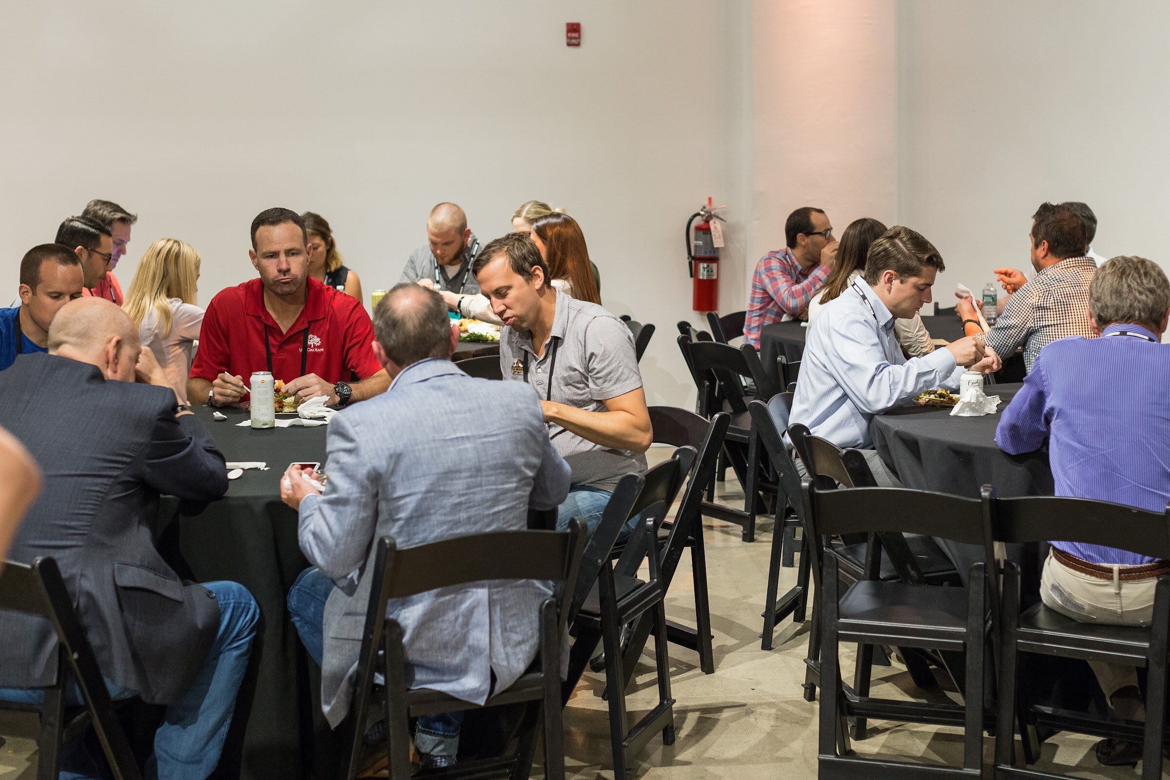 Attendees sit at black-draped round tables eating lunch with cans of beverage on the tables.