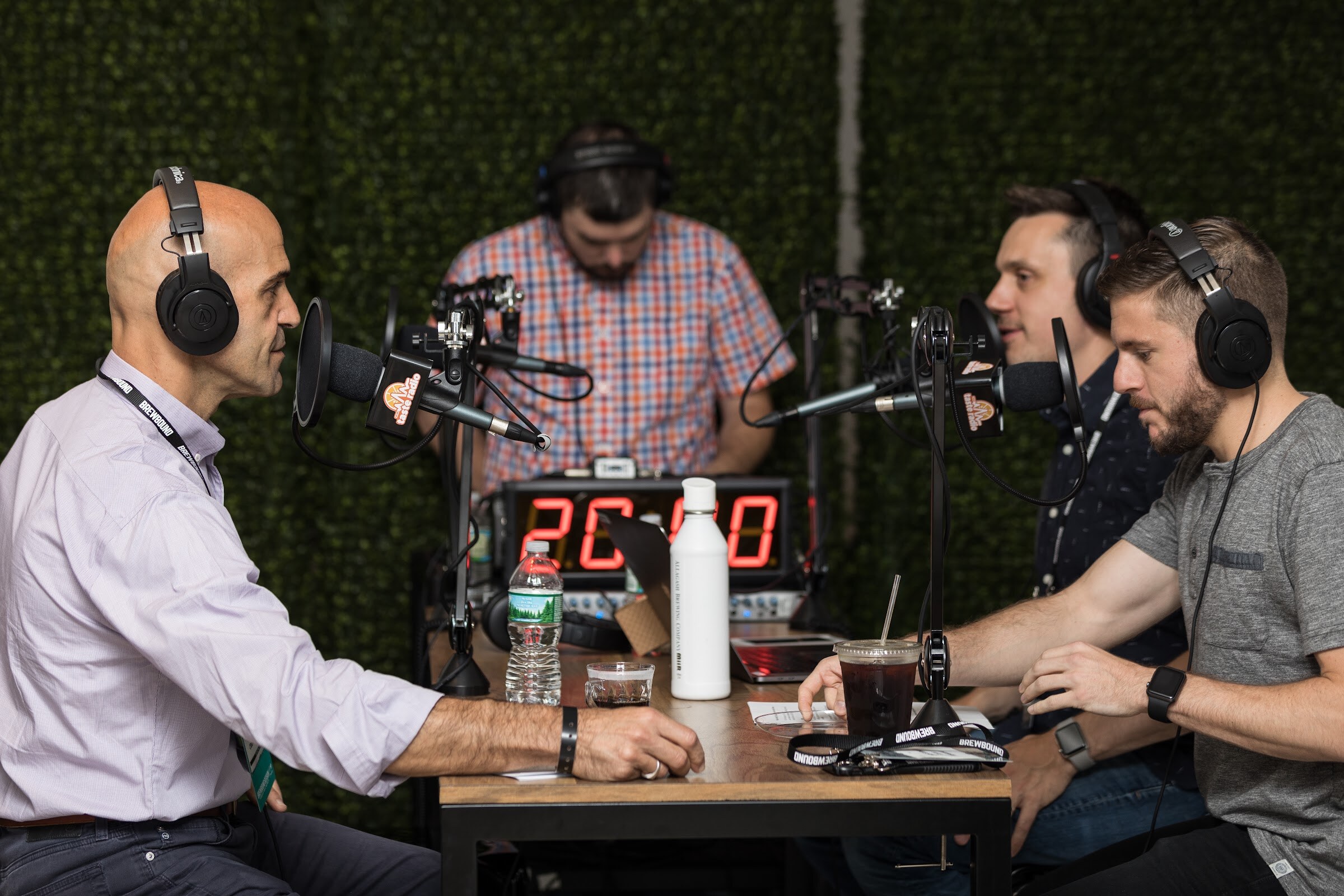 Three men in headphones sit at a wooden table with microphones carrying the Taste Radio logo and a red countdown timer while a fourth person works the audio board behind them against a hedge wall.