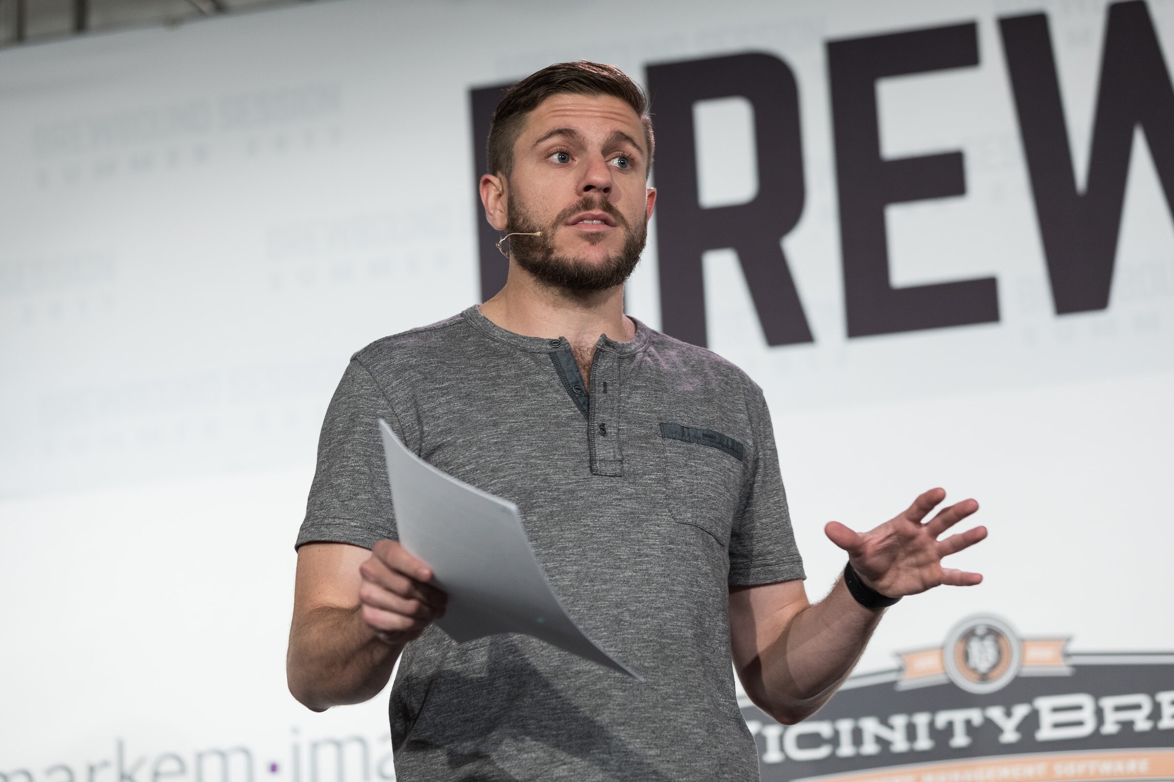 A bearded man in a gray henley with a headset microphone holds a sheet of paper and gestures in front of a large Brewbound Session backdrop with a Vicinity Brew logo.