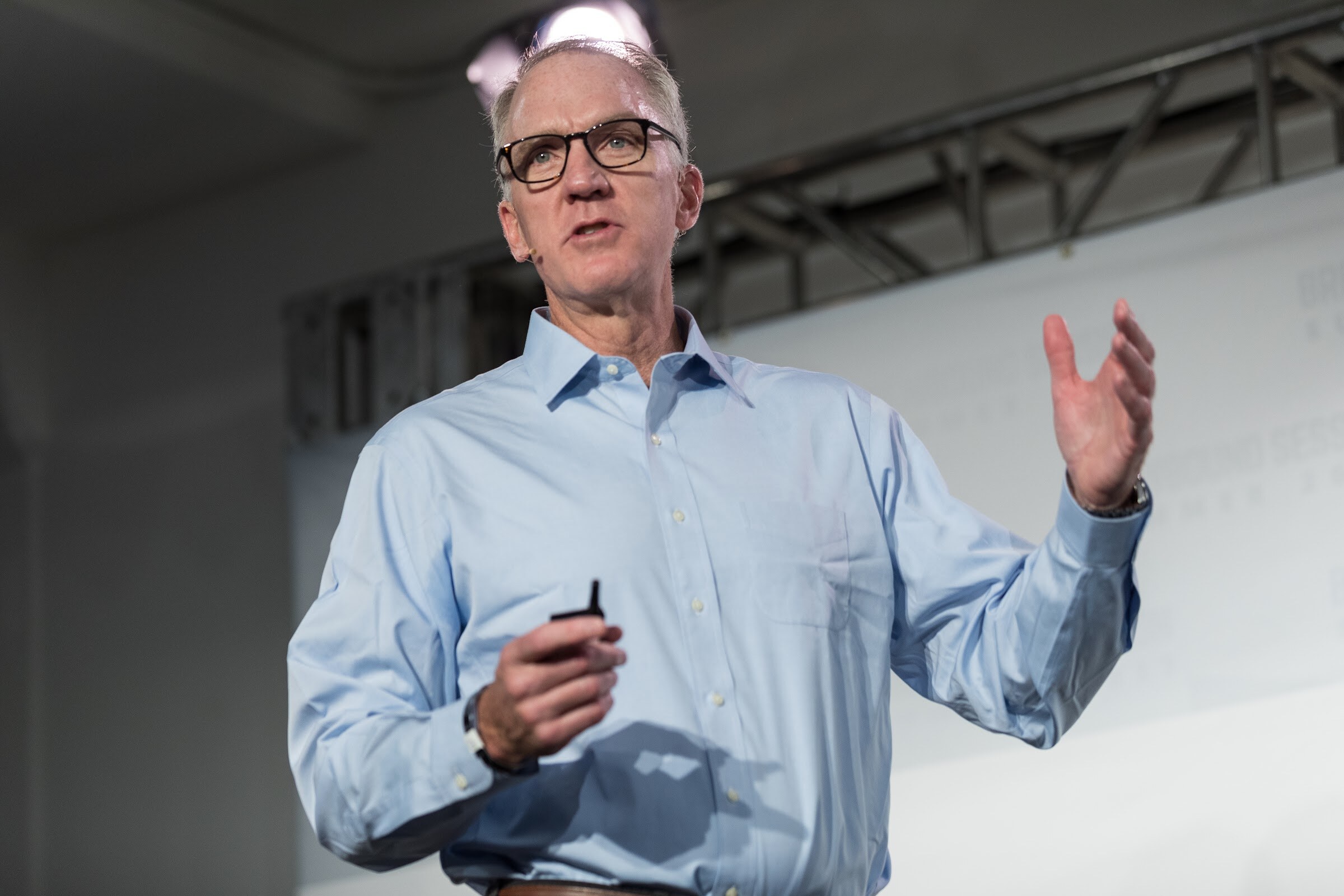 A man in a light blue shirt and dark glasses holds a clicker and raises one hand while speaking on stage.