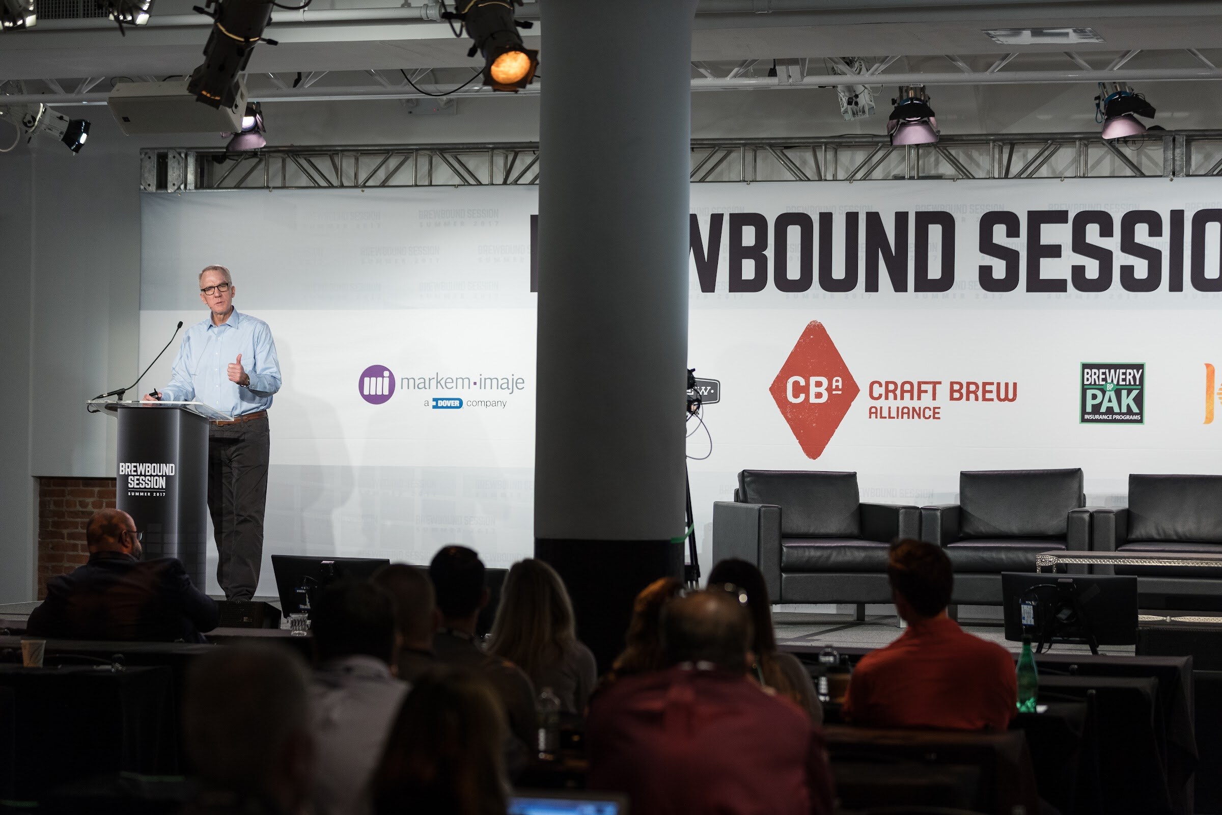 A man in a light blue shirt speaks at a black Brewbound Session podium on a stage with Markem-Imaje, Craft Brew Alliance and Brewery Pak signs, with audience heads in the foreground.