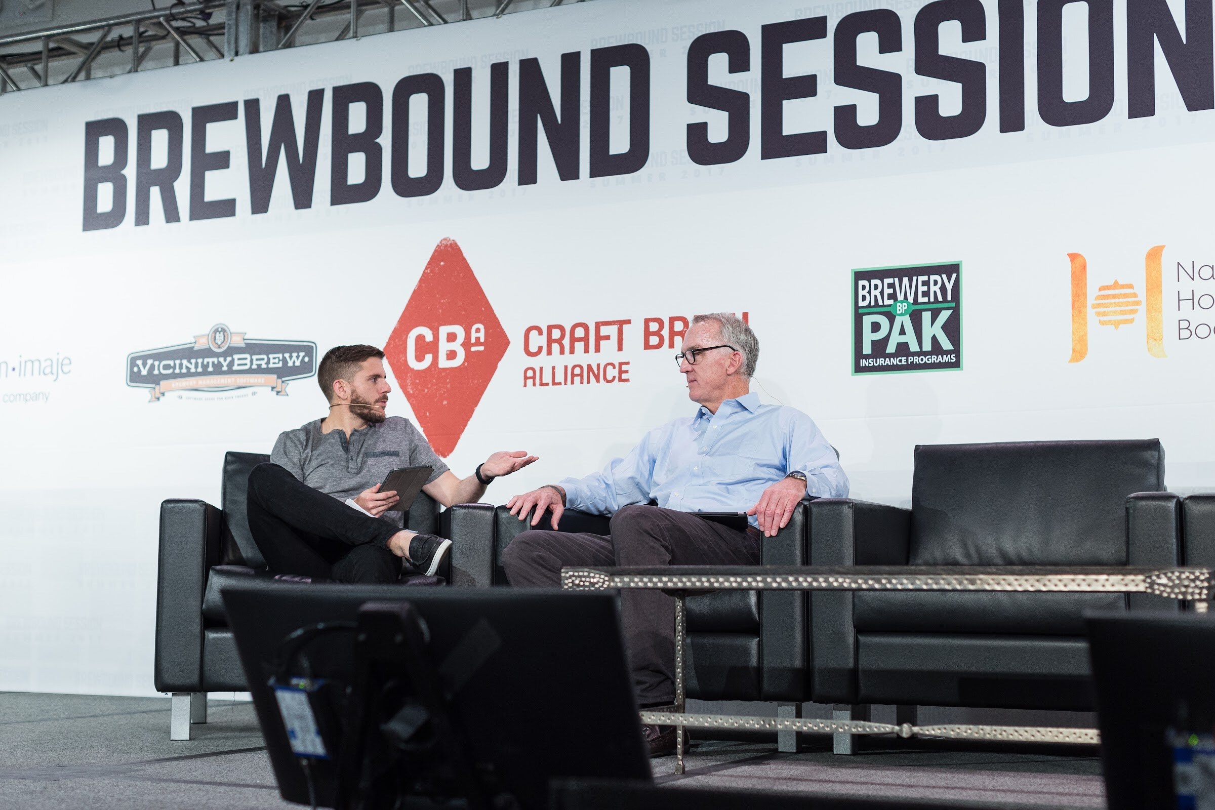 A host in a gray henley holding a tablet and a man in a light blue shirt talk on black couches in front of Vicinity Brew, Craft Brew Alliance and Brewery Pak signs.