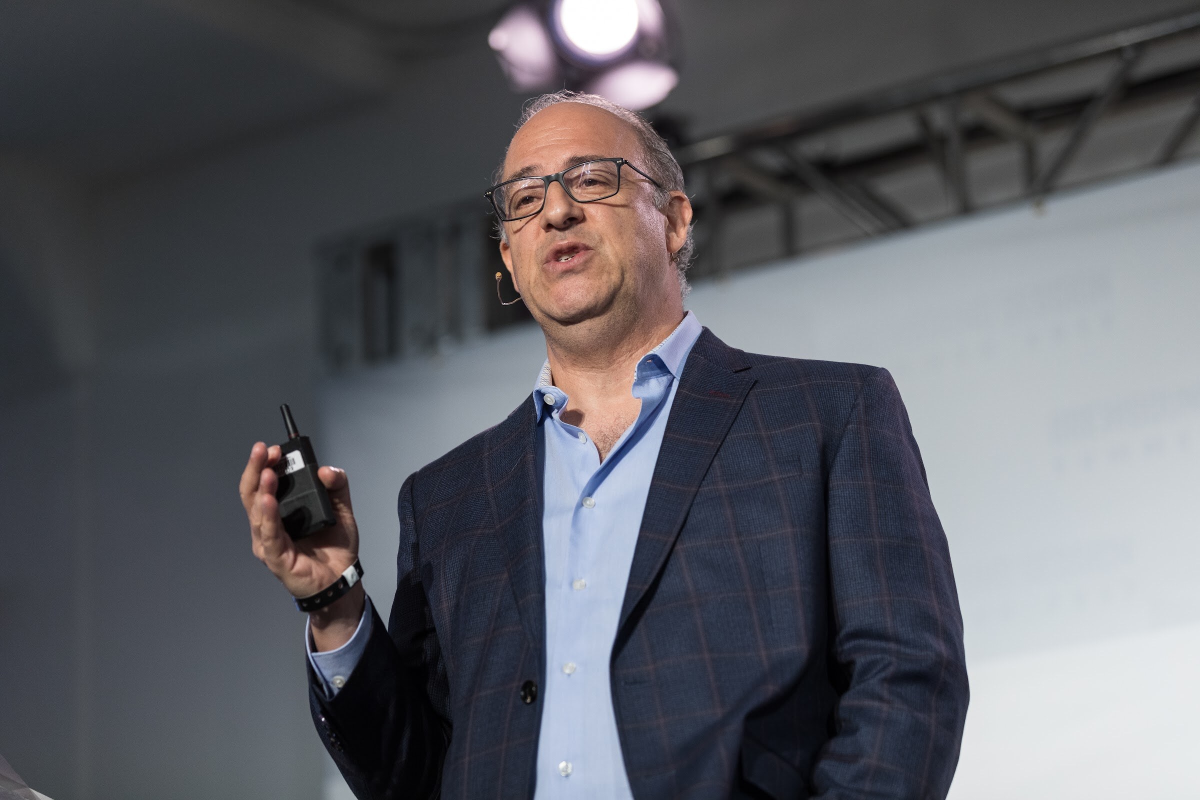 A man with glasses and a navy plaid blazer holds a presentation clicker and speaks under stage lights.