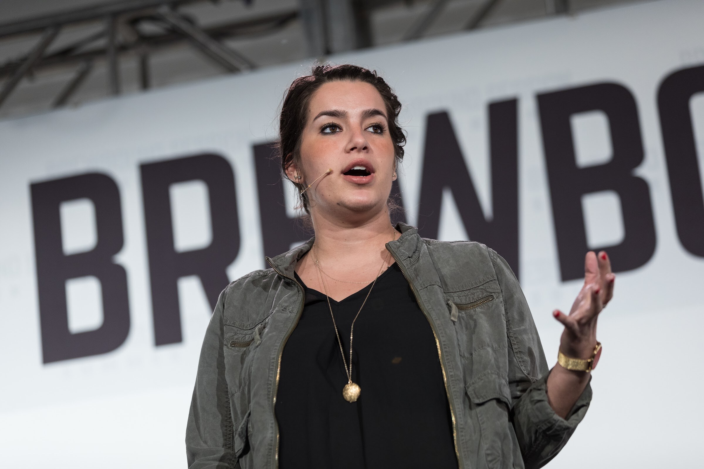 A woman in an olive utility jacket and black top speaks into a headset microphone with a raised hand in front of big dark Brewbound lettering.