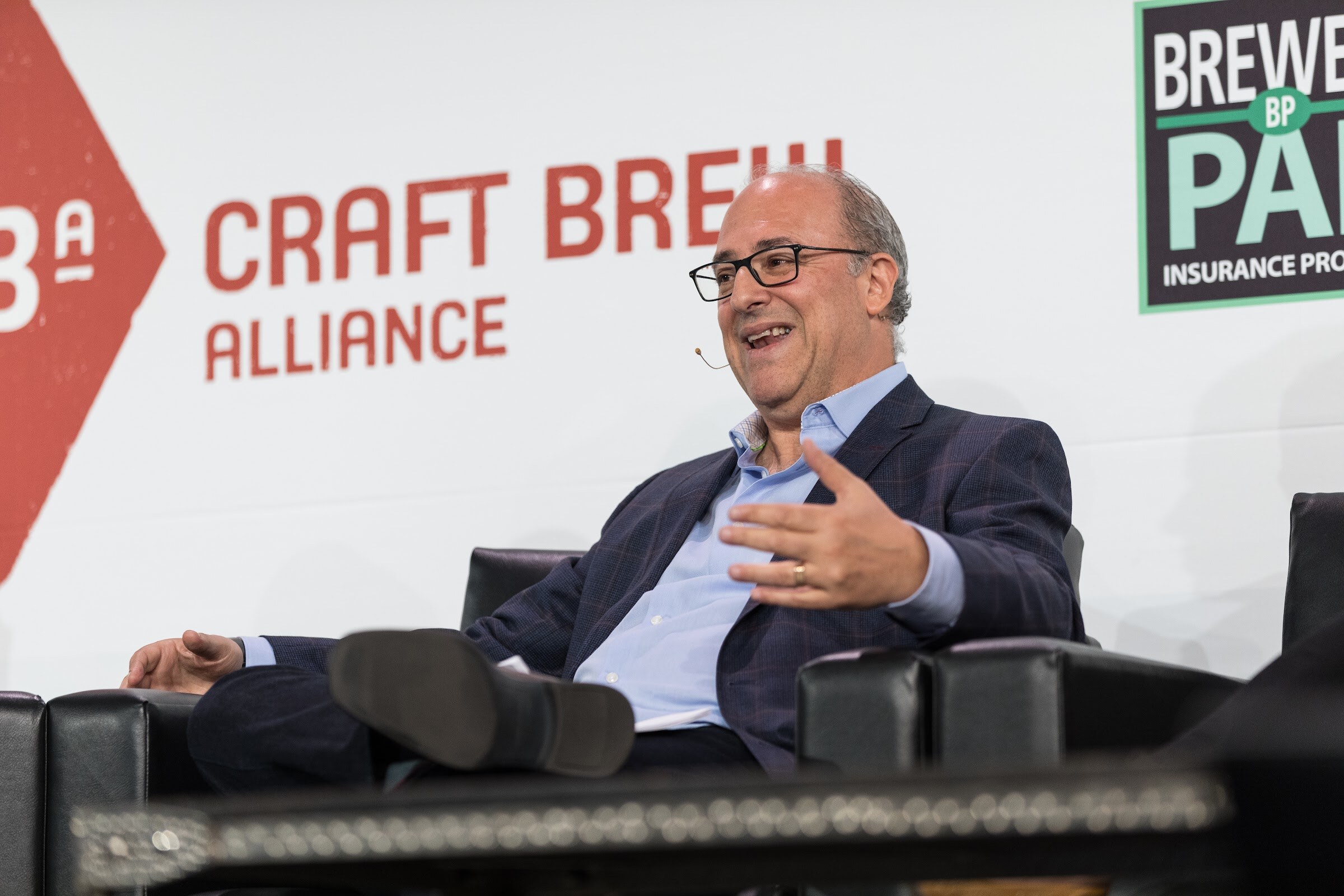 A man in a navy blazer and glasses laughs and gestures from a black leather armchair in front of Craft Brew Alliance and Brewery Pak signs.