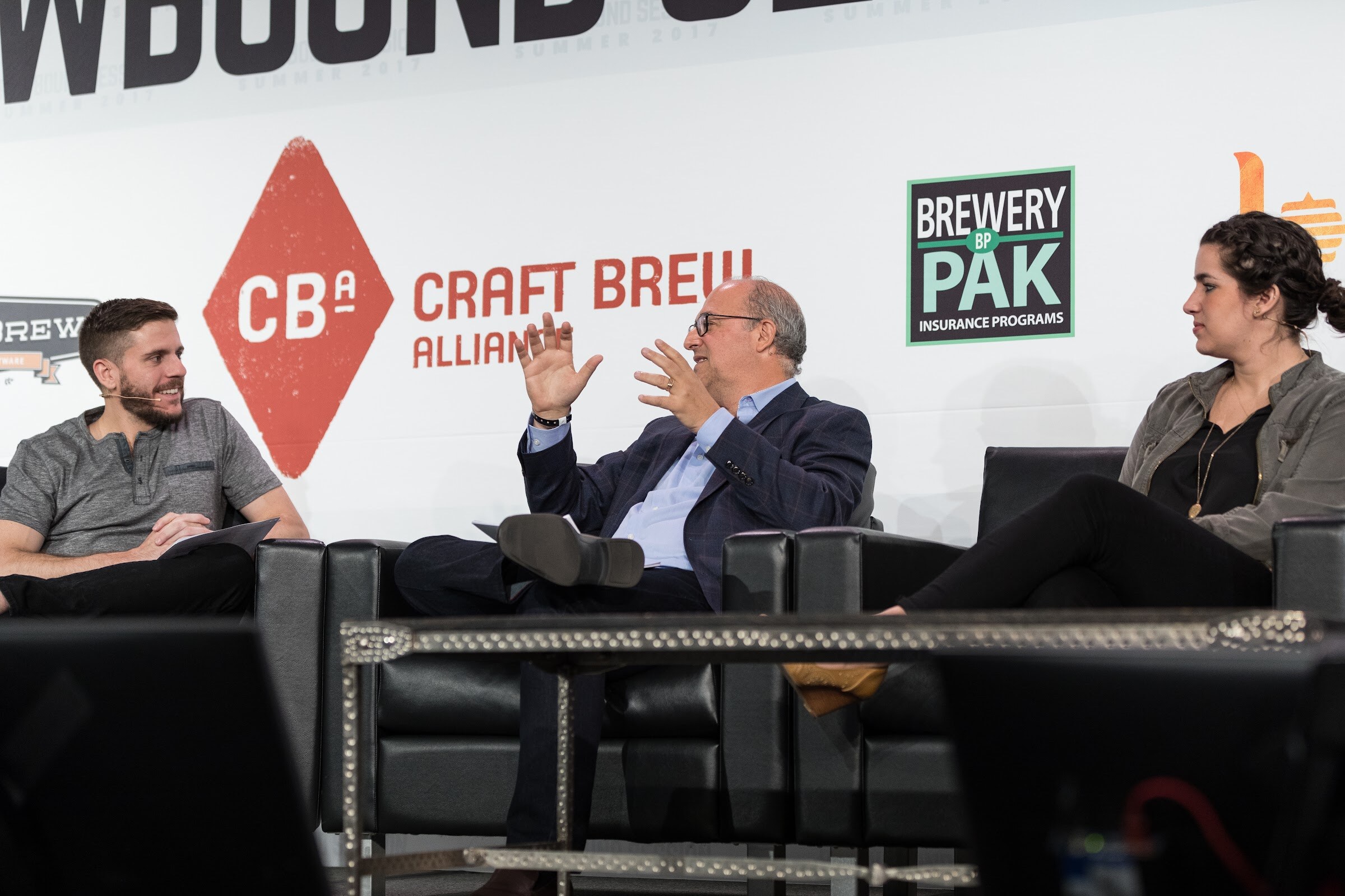 A bearded host in a gray henley, a man in a navy blazer and a woman in an olive jacket sit on black leather couches in front of Craft Brew Alliance and Brewery Pak signs.