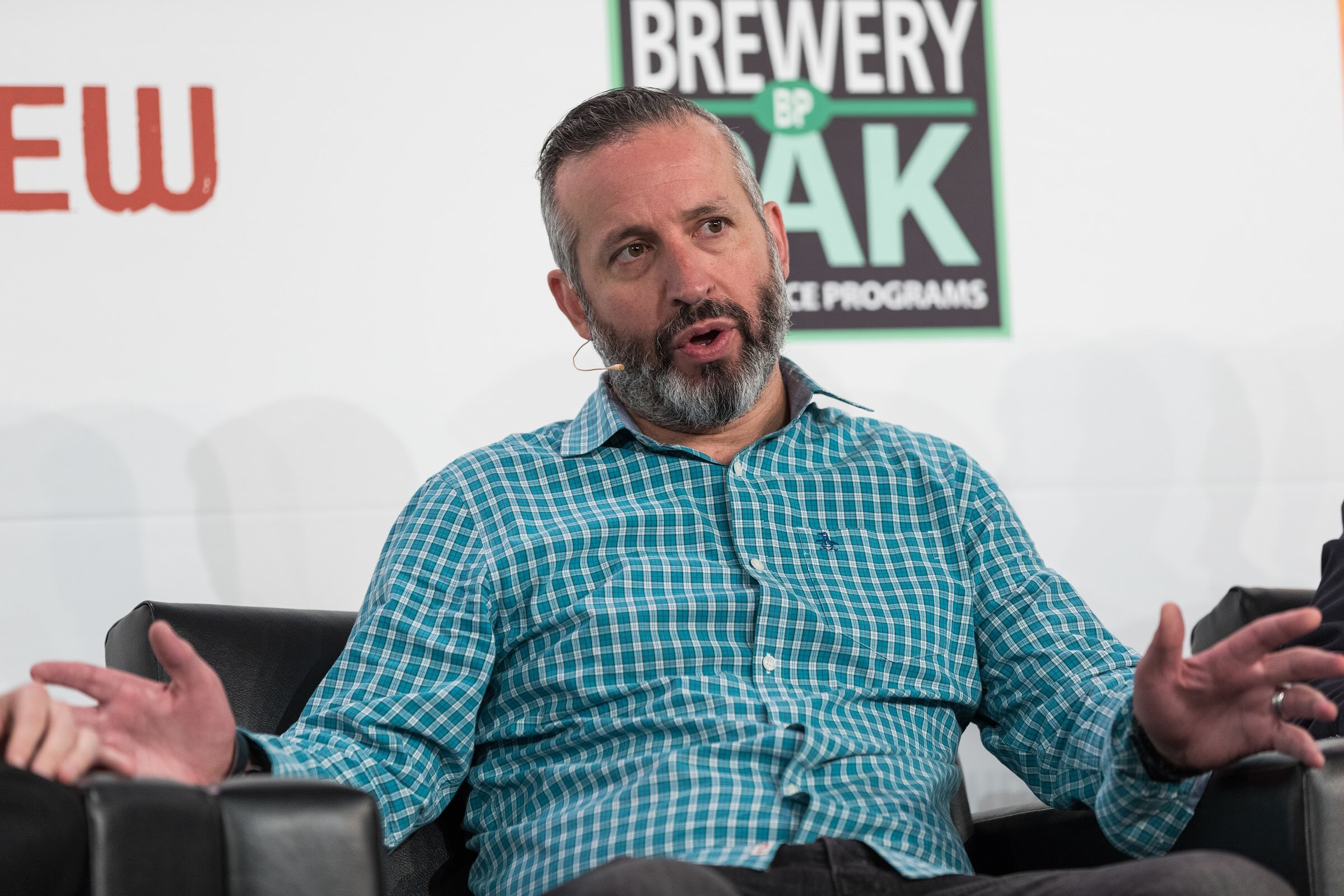 A bearded man in a teal checked shirt gestures with both hands from a black armchair in front of a Brewery Pak sign.