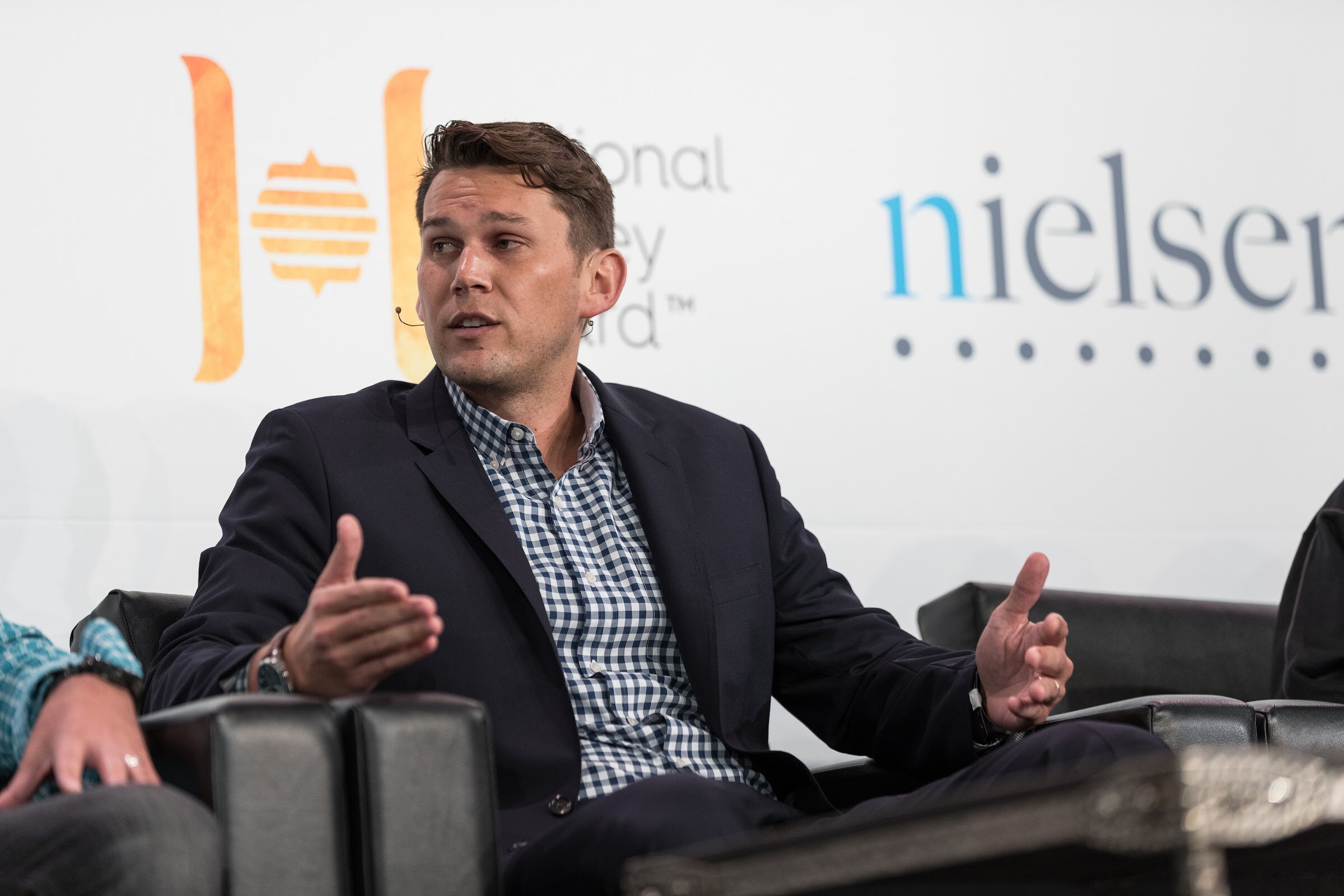 A man in a navy blazer and checked shirt gestures from a black armchair in front of National Honey Board and Nielsen signs.