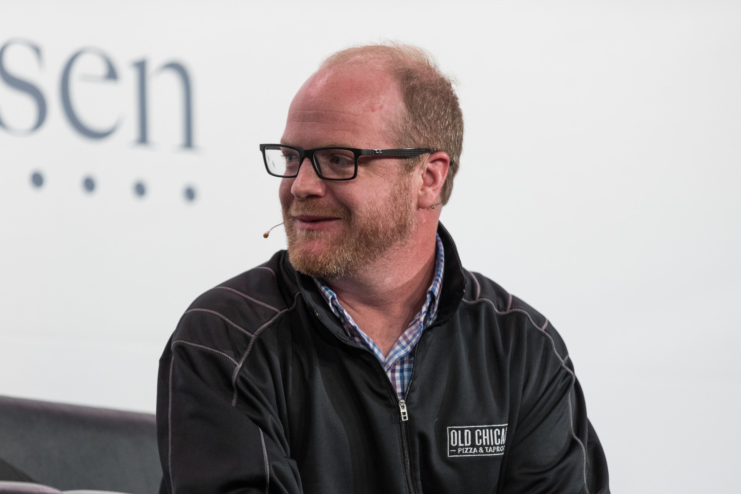 A bearded man with black glasses in a black zip jacket reading Old Chicago smiles and looks to his left in front of a Nielsen sign.