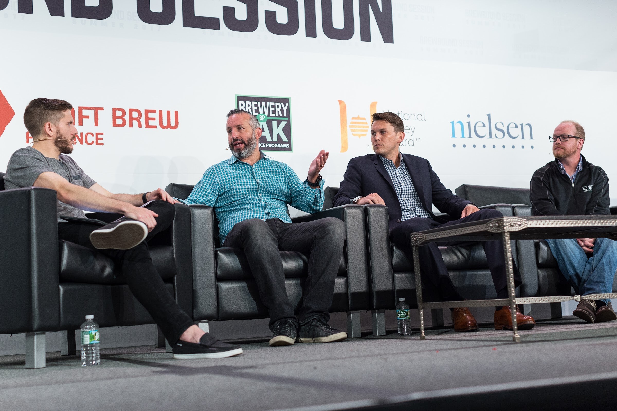 Four men sit on black leather couches on stage in front of Craft Brew Alliance, Brewery Pak, National Honey Board and Nielsen signs while one gestures mid-sentence.
