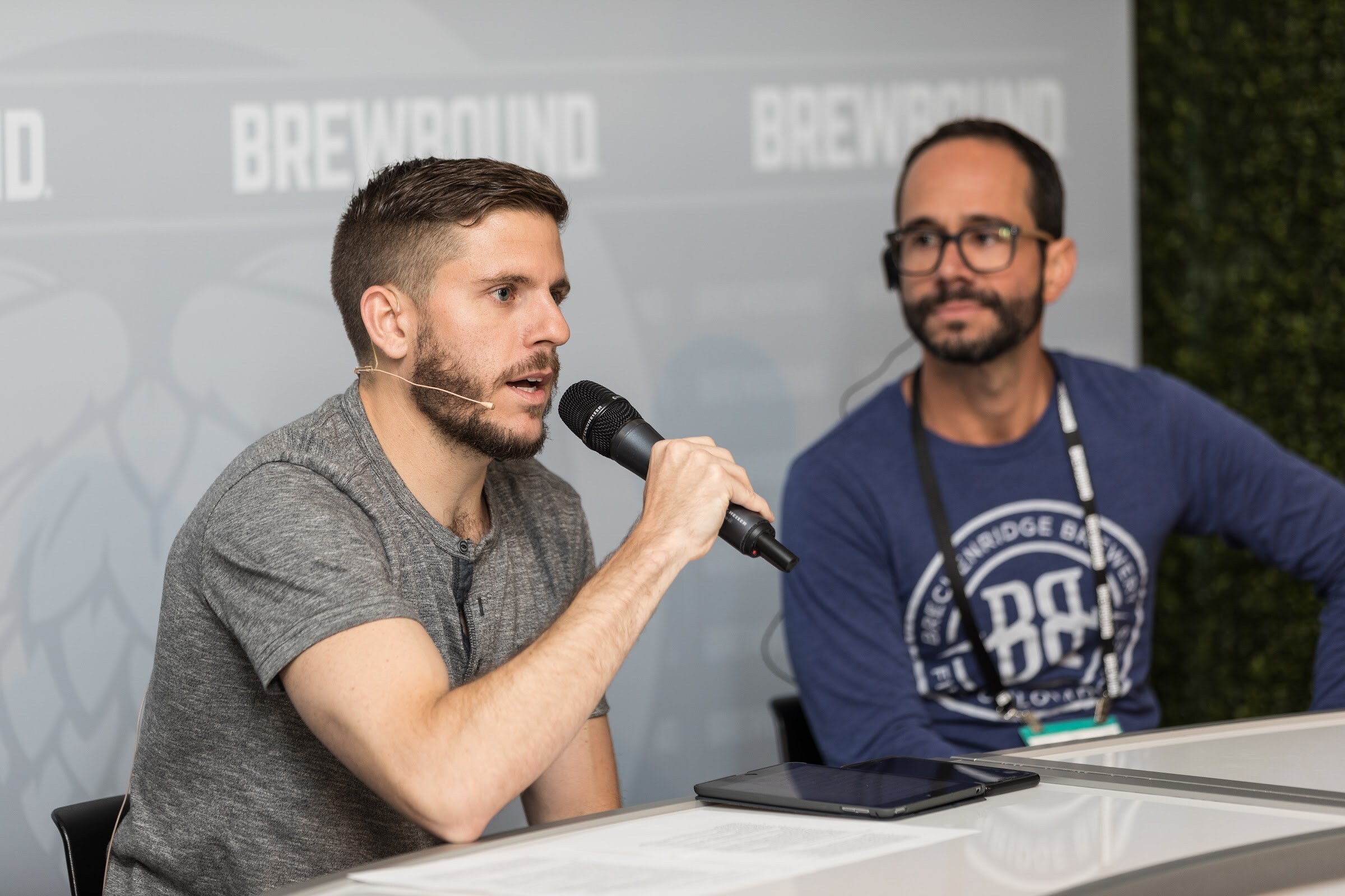 A man in a gray shirt speaks into a handheld microphone while a bearded man with glasses in a navy shirt with a Breckenridge Brewery logo sits beside him in front of a Brewbound backdrop.