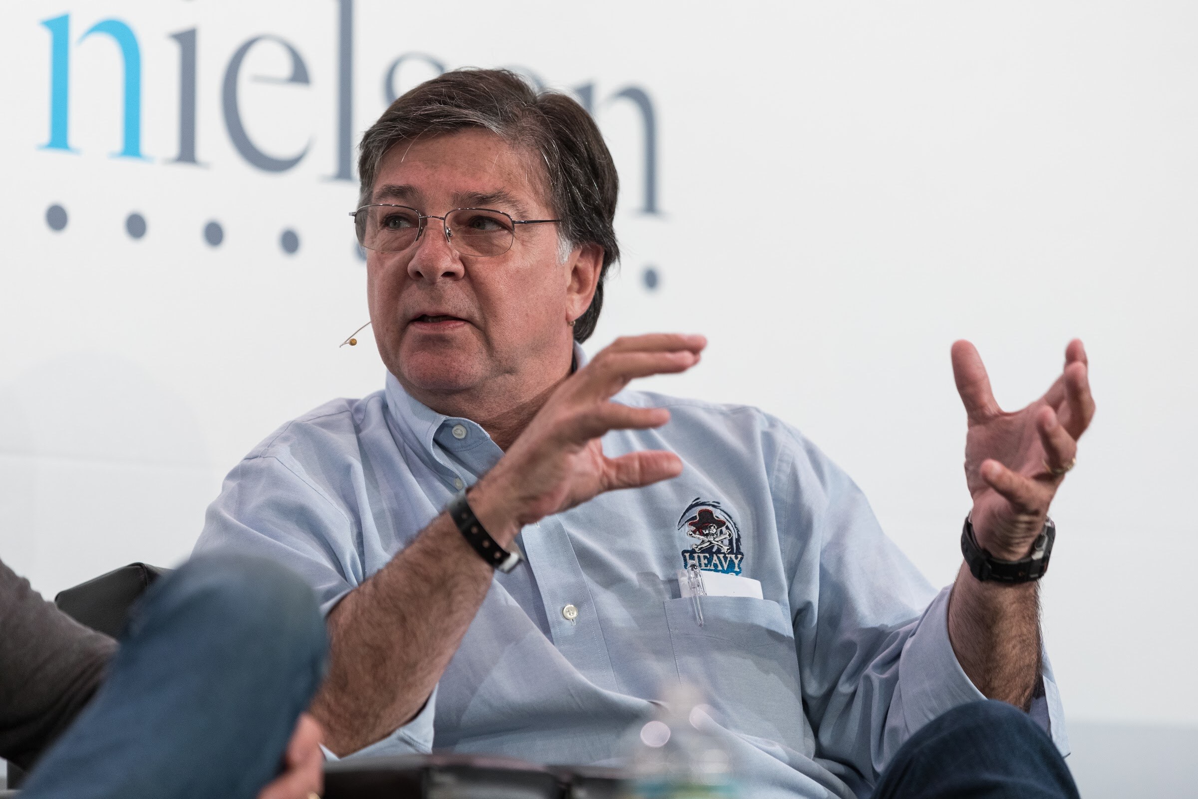 A gray-haired man with glasses in a light blue shirt with a Heavy Seas logo gestures with both hands in front of a Nielsen sign.