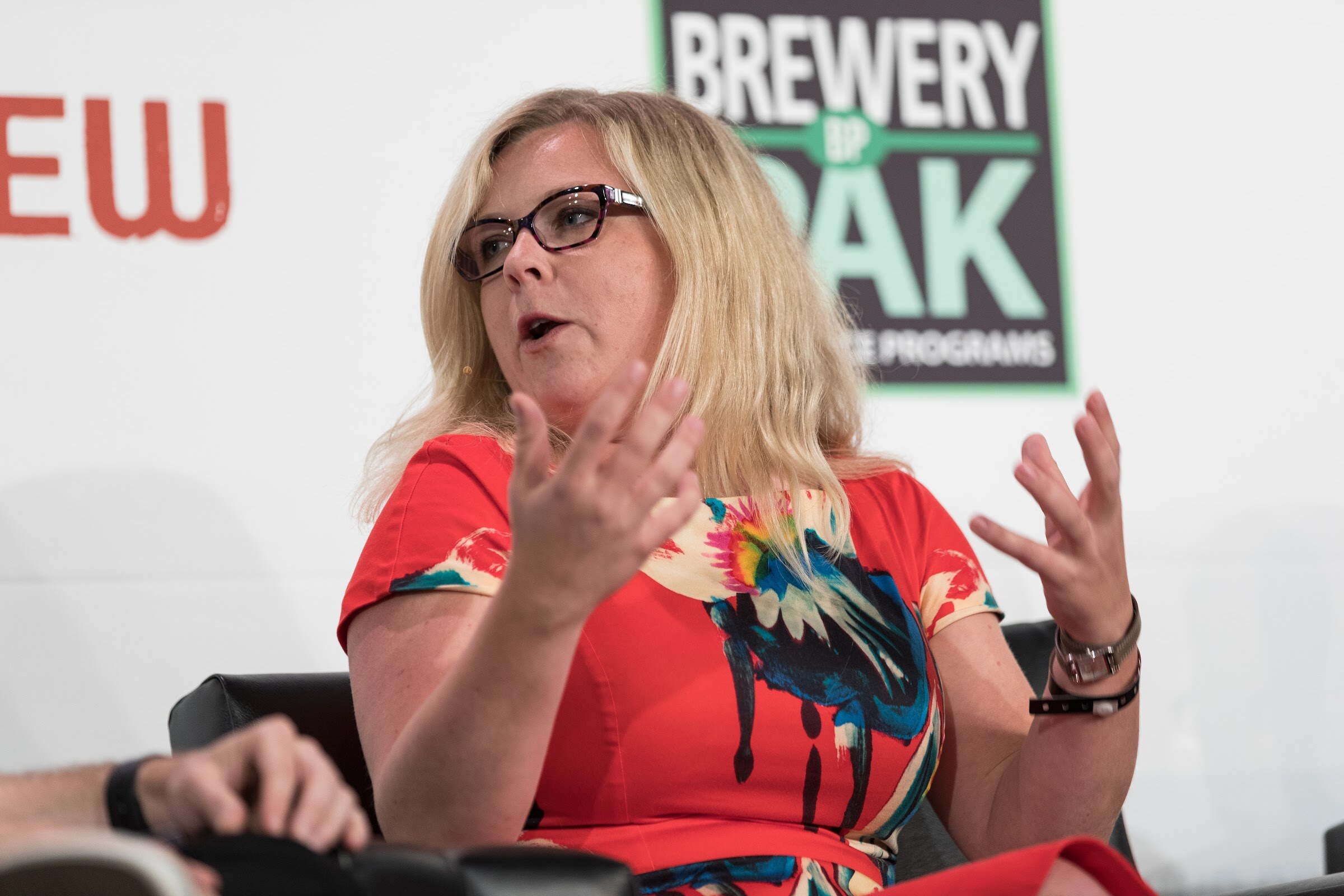 A woman with glasses and blonde hair in a red floral dress gestures with both hands in front of a Brewery Pak sign.