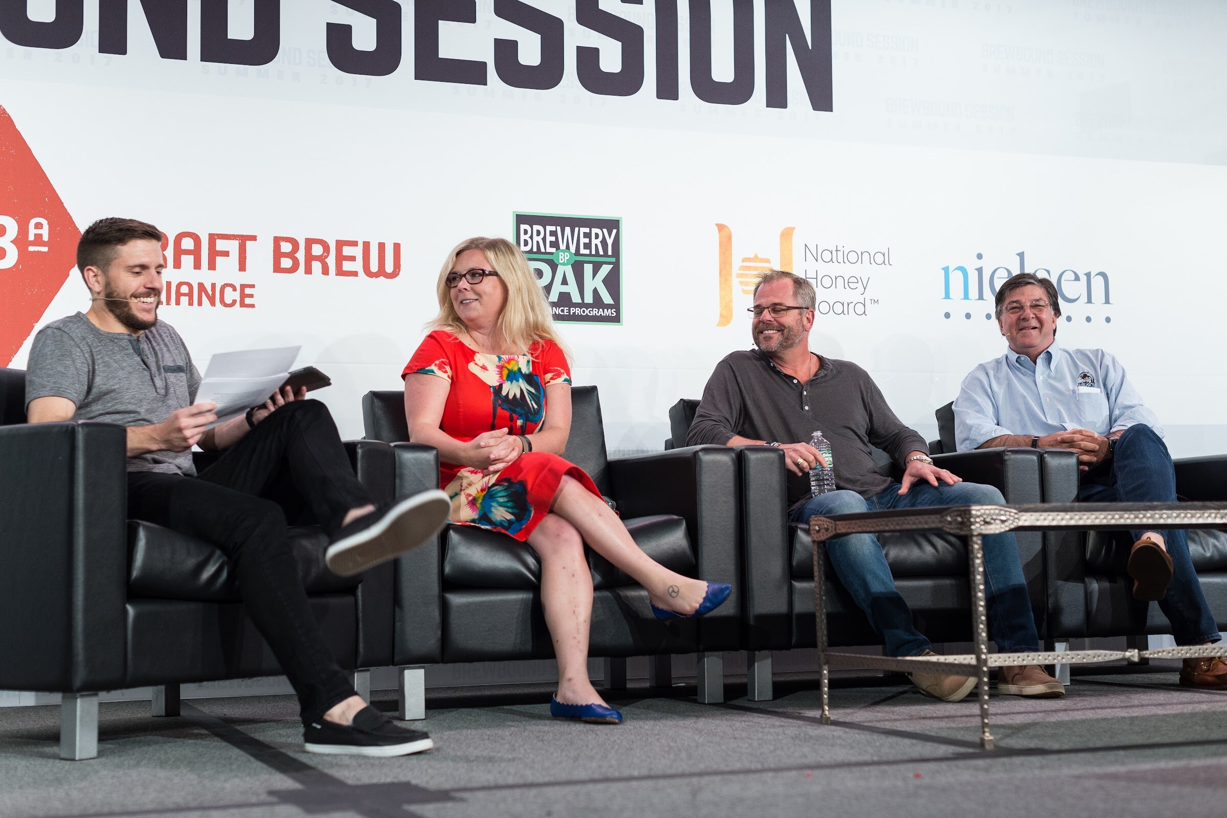 A host holding papers and tablet and three panelists sit in a row on black leather couches in front of Craft Brew Alliance, Brewery Pak, National Honey Board and Nielsen signs.