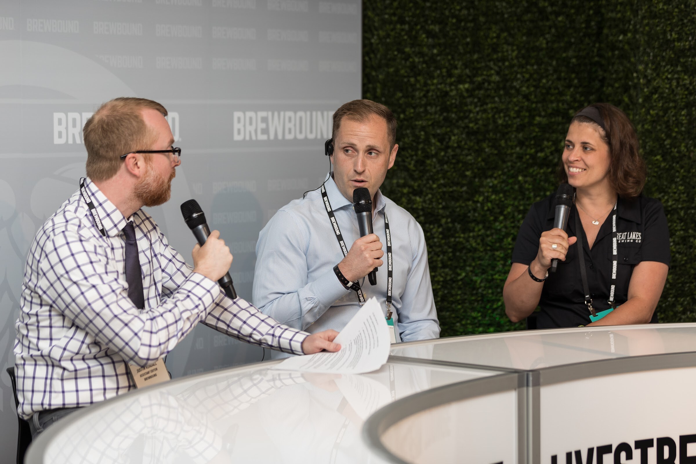 A man in a checked shirt and tie holding papers sits with two guests holding microphones at a white curved desk labeled Livestream, with a hedge wall behind them; one guest wears a Great Lakes Brewing shirt.